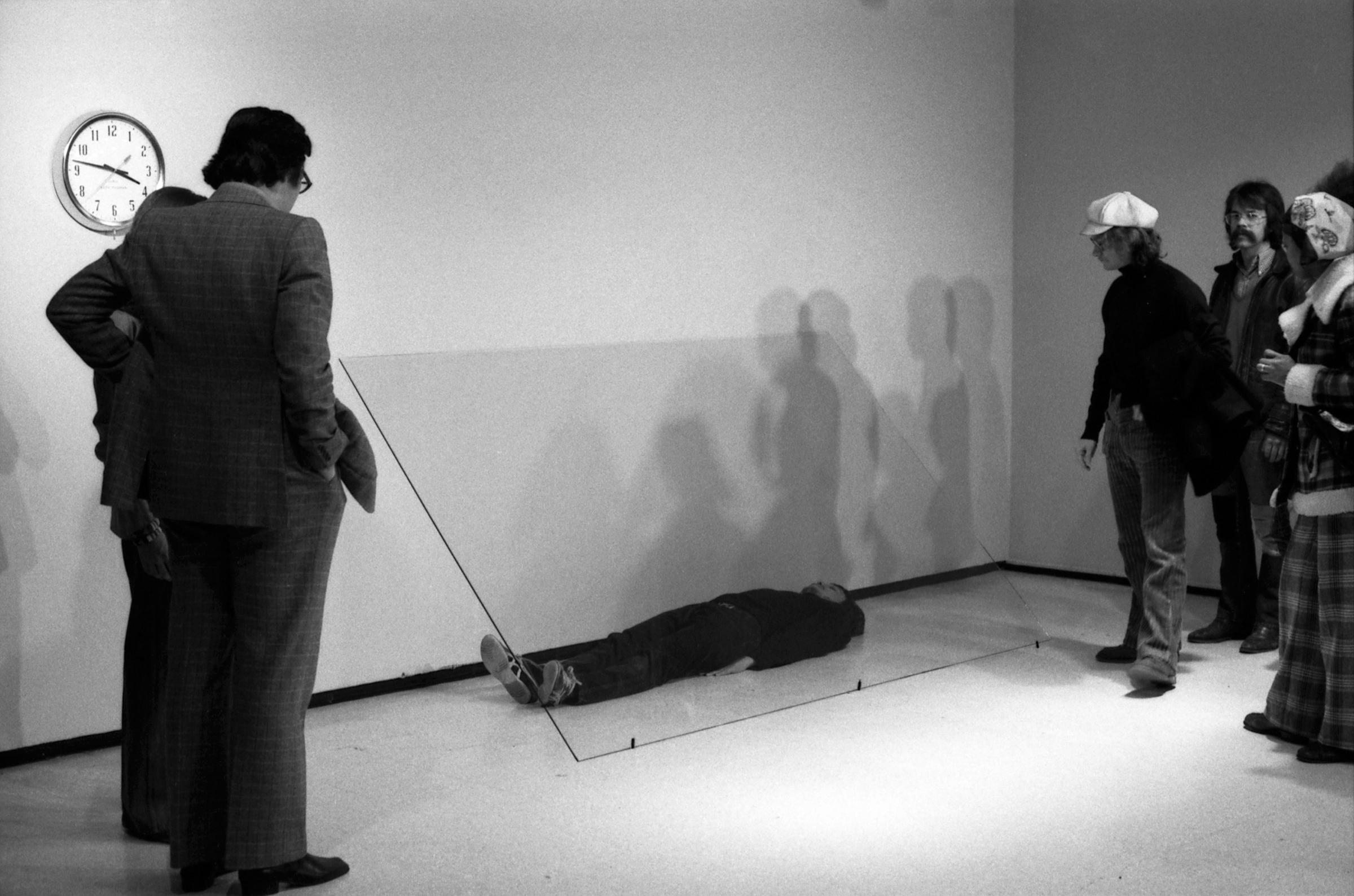 A group of museum visitors observe a man lying on the floor underneath a sheet of glass.