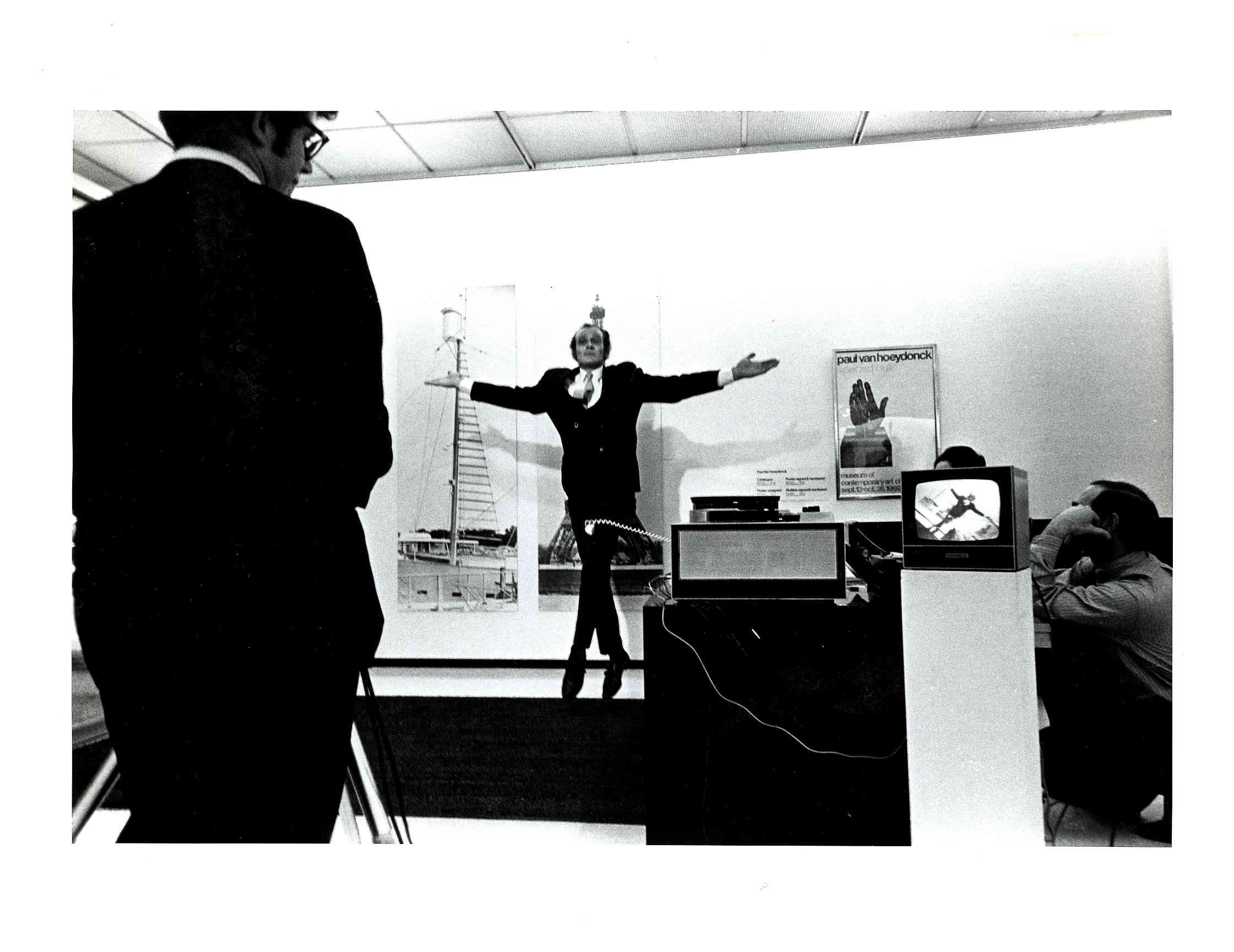 Black and white photo of a man jumping inside a gallery-like space. His movement is simultaneously projected on a small television.