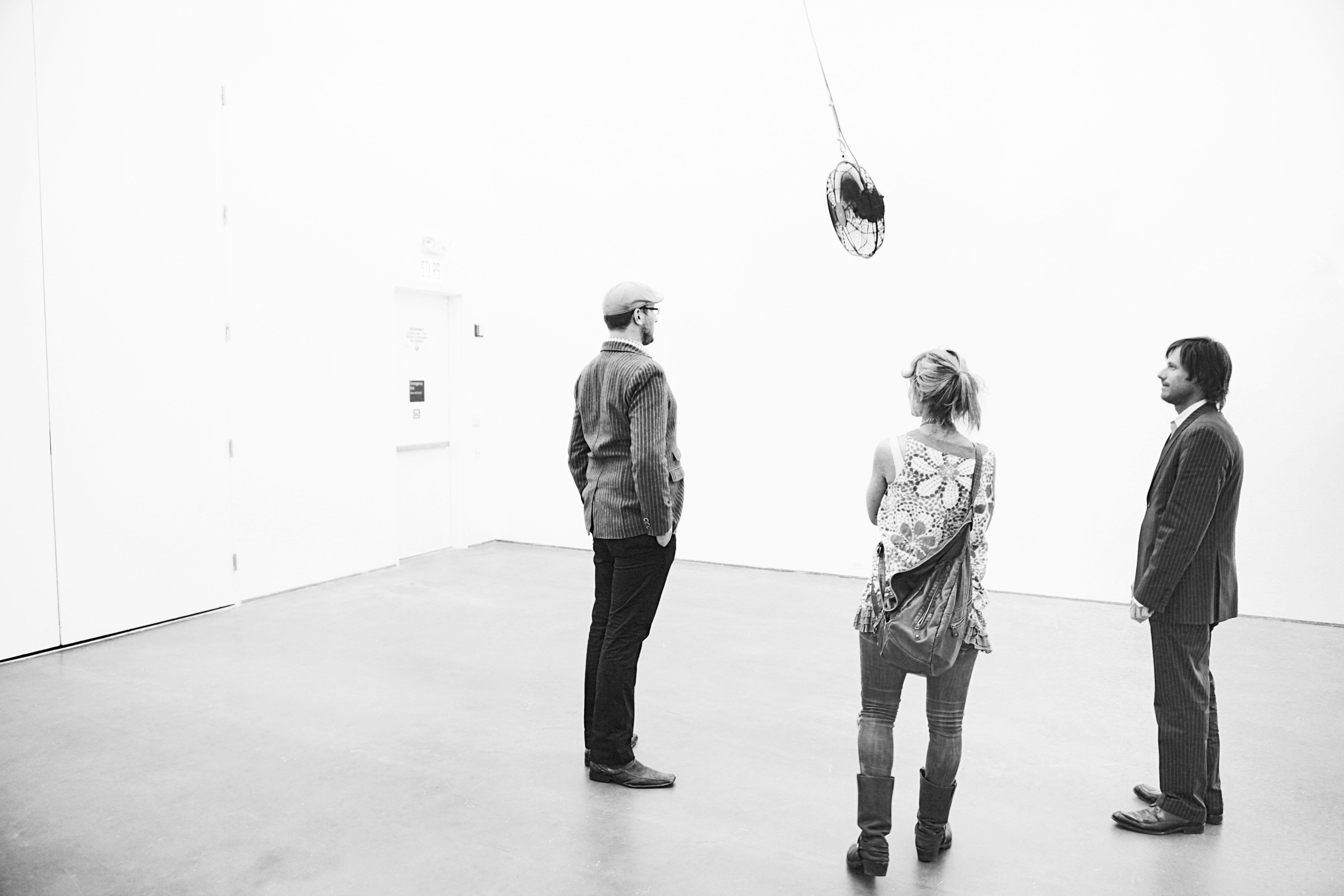 Three people in a black-and-white photograph look at a ceiling fan hanging from the ceiling.