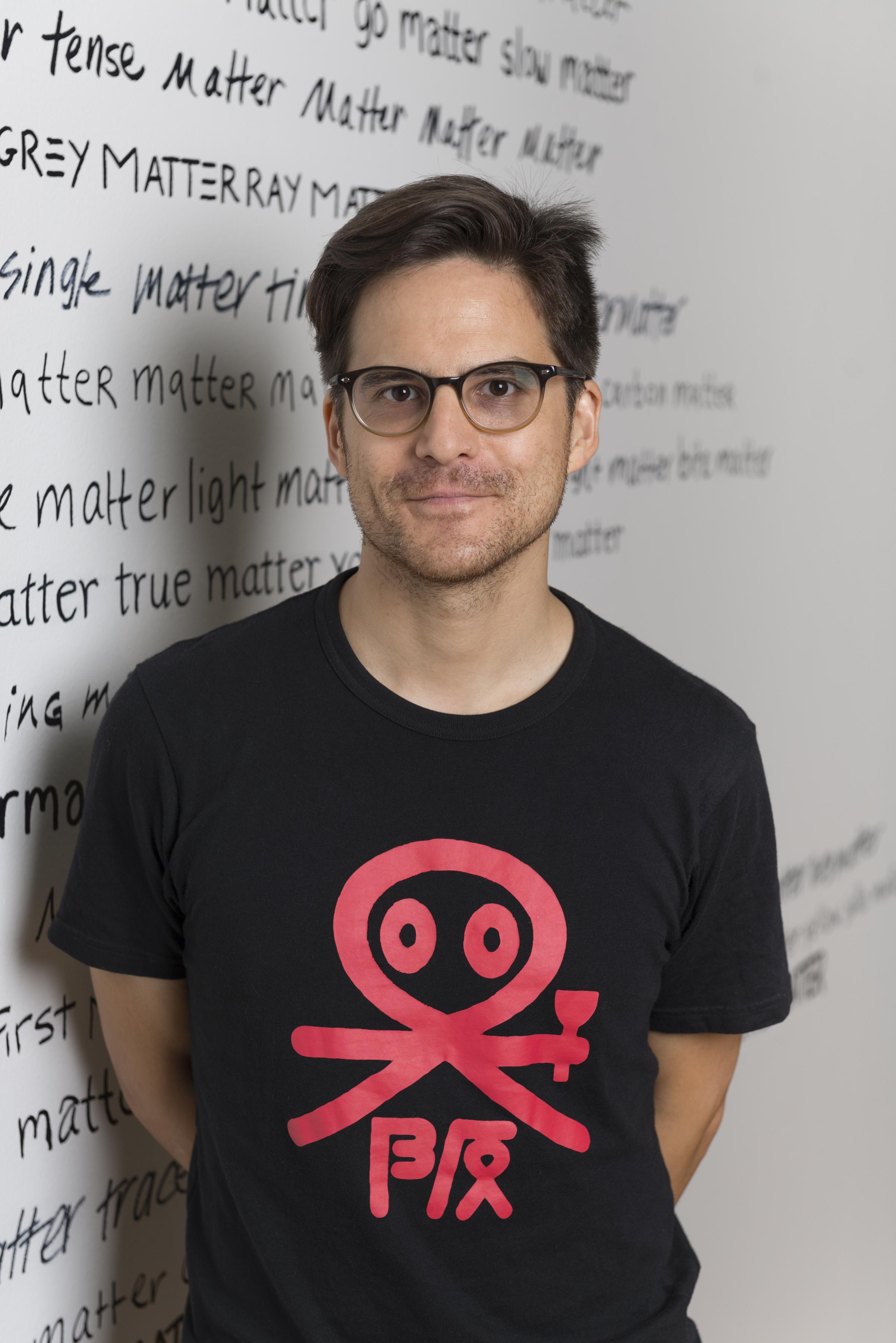 A man wearing glasses and a black t-shirt stands in front of a wall with text written in various scripts repeatedly saying "matter" in addition to adjectives such as "single," "true," and "slow."