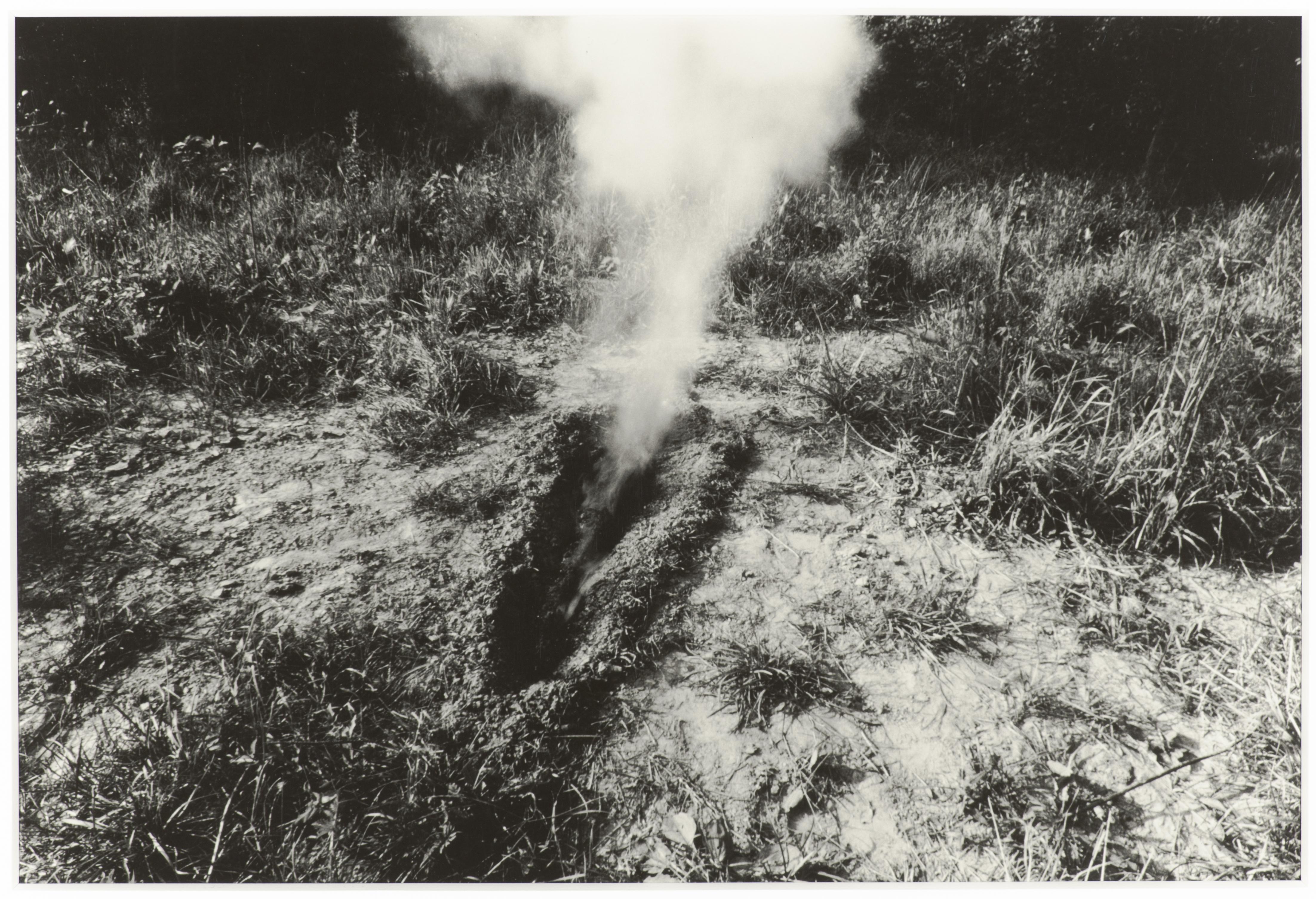 A black-and-white photo captures smoke billowing from a body-sized trench in a clearing with weeds and trees in the horizon.