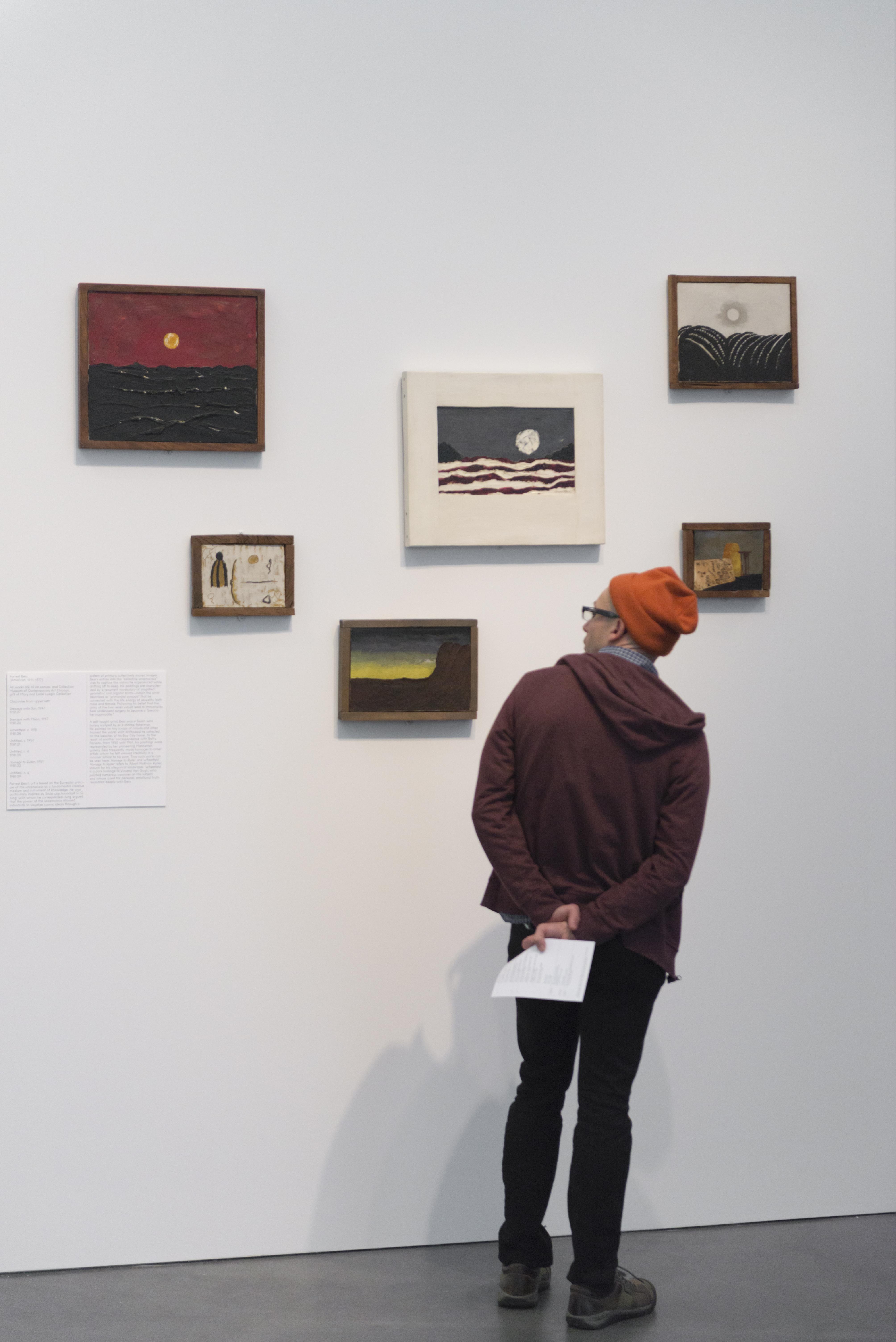 A person wearing a hoodie and beanie faces six small and medium sized paintings on a gallery wall.