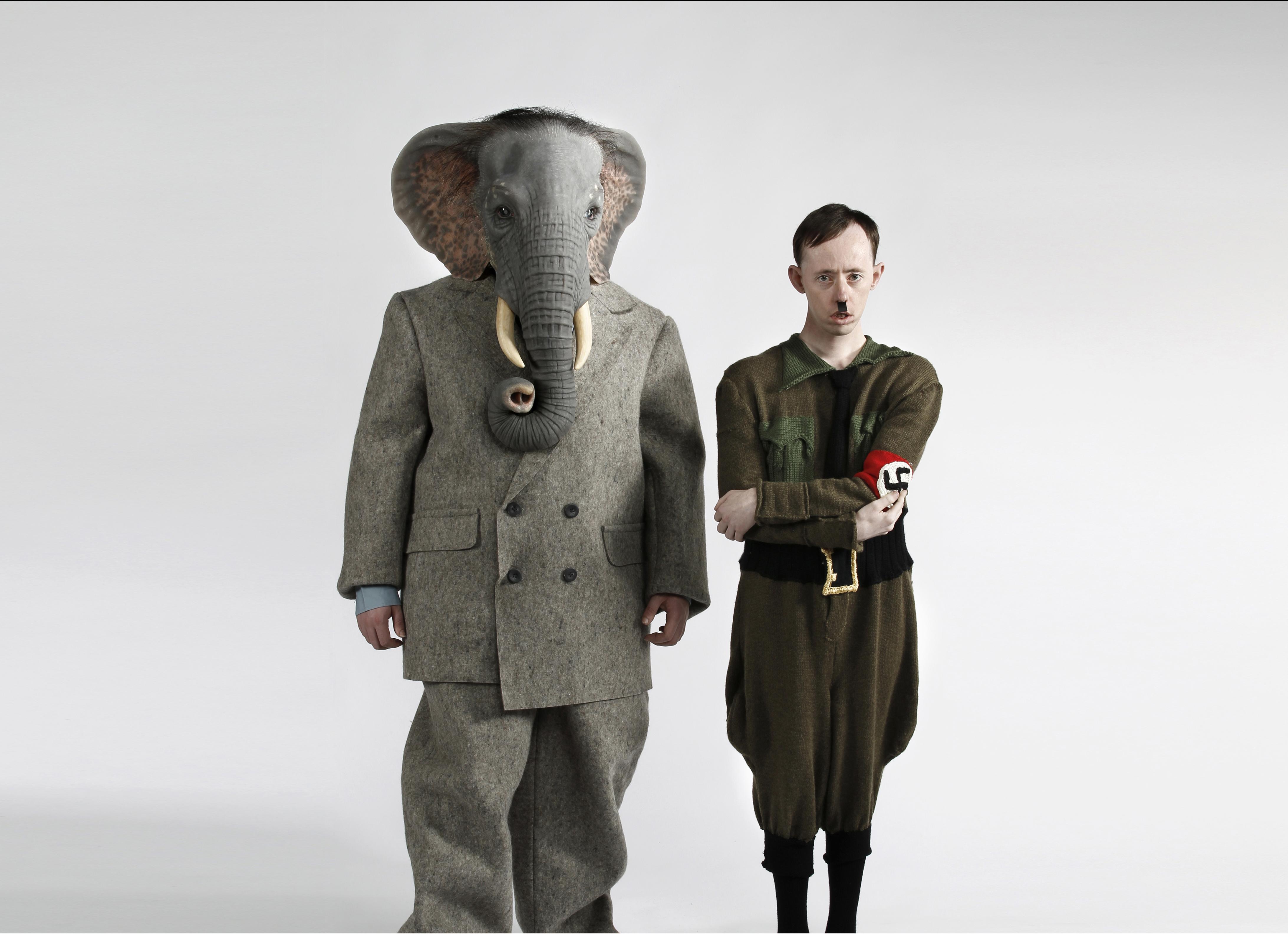 A person with a head like an elephant wears a gray business suit and stands beside a person in a nazi uniform who resembles Adolf Hitler.