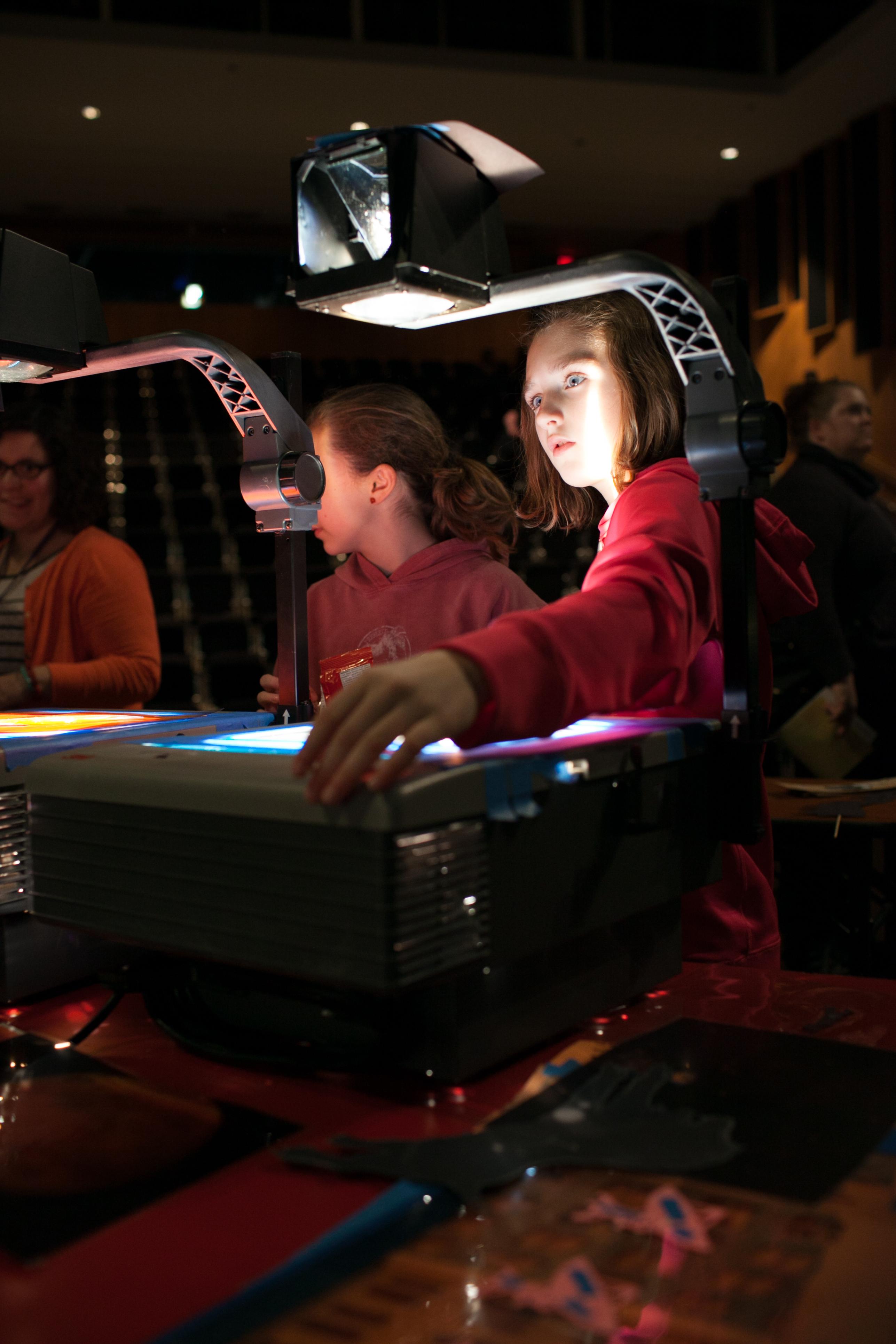 Two children in hooded sweatshirts stand behind overhead projectors, their faces illuminated by the light of the machines.