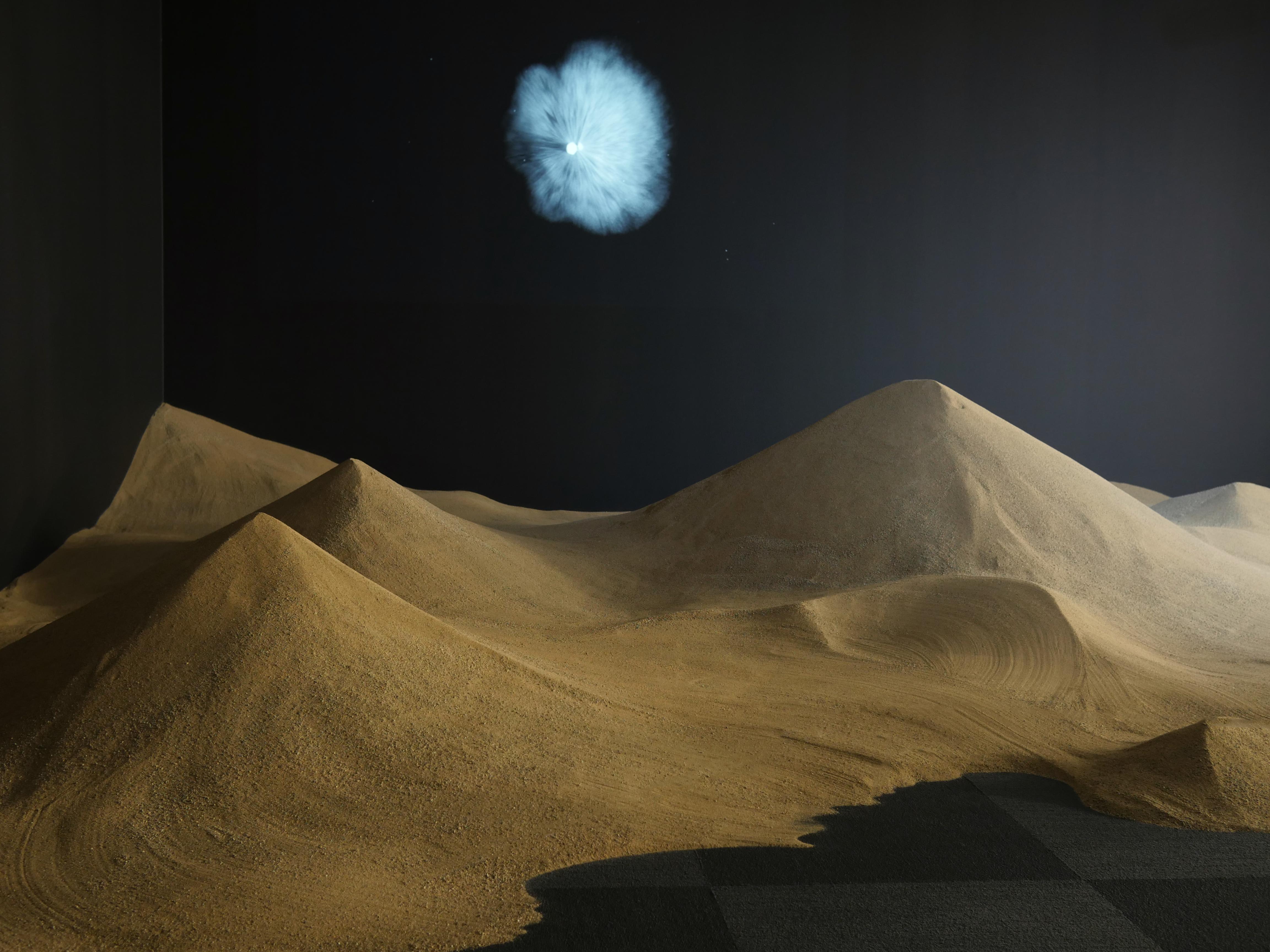 Dimly lit sand dunes stand out against a dark black background. An irregularly shaped light formation is projected on the wall above the dunes like a moon rising in the night sky.