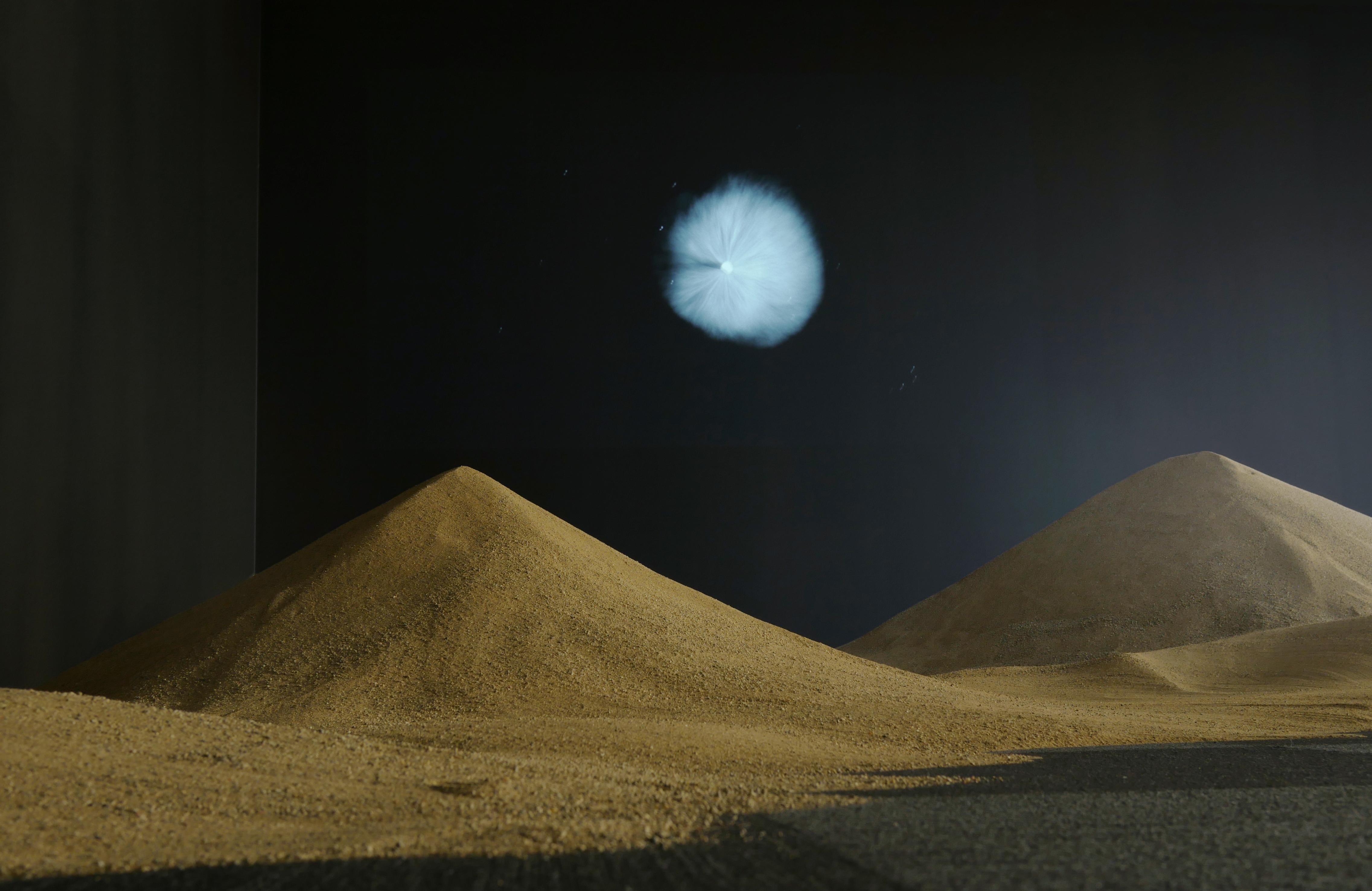 Two dunes of sand are dimly lit in a dark room. Above them is a video projection of a light or some astronomical event.