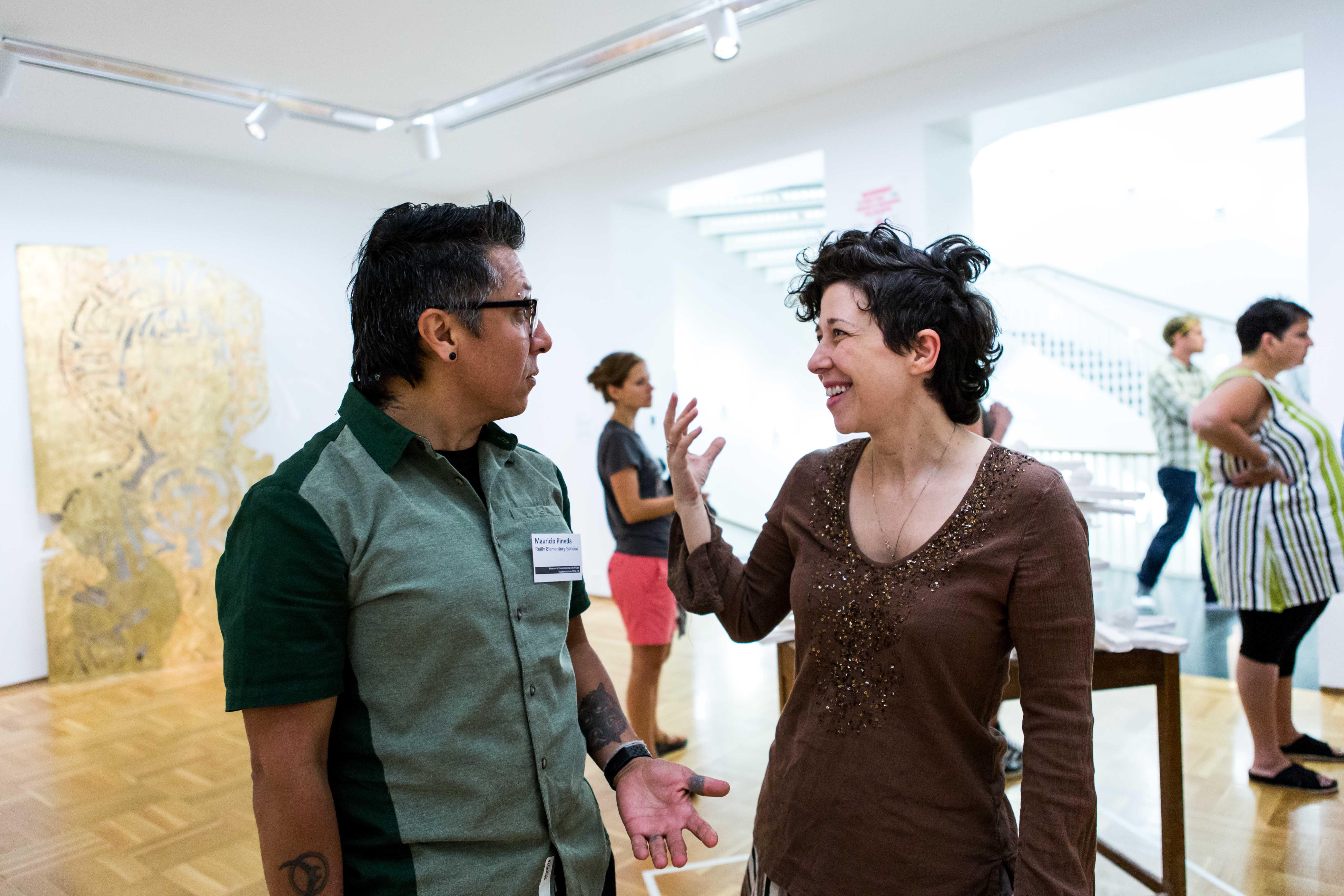 A man and a woman engage in a lively conversation in a gallery; an artwork resting against the wall and three out-of-focus museum visitors are in the background.