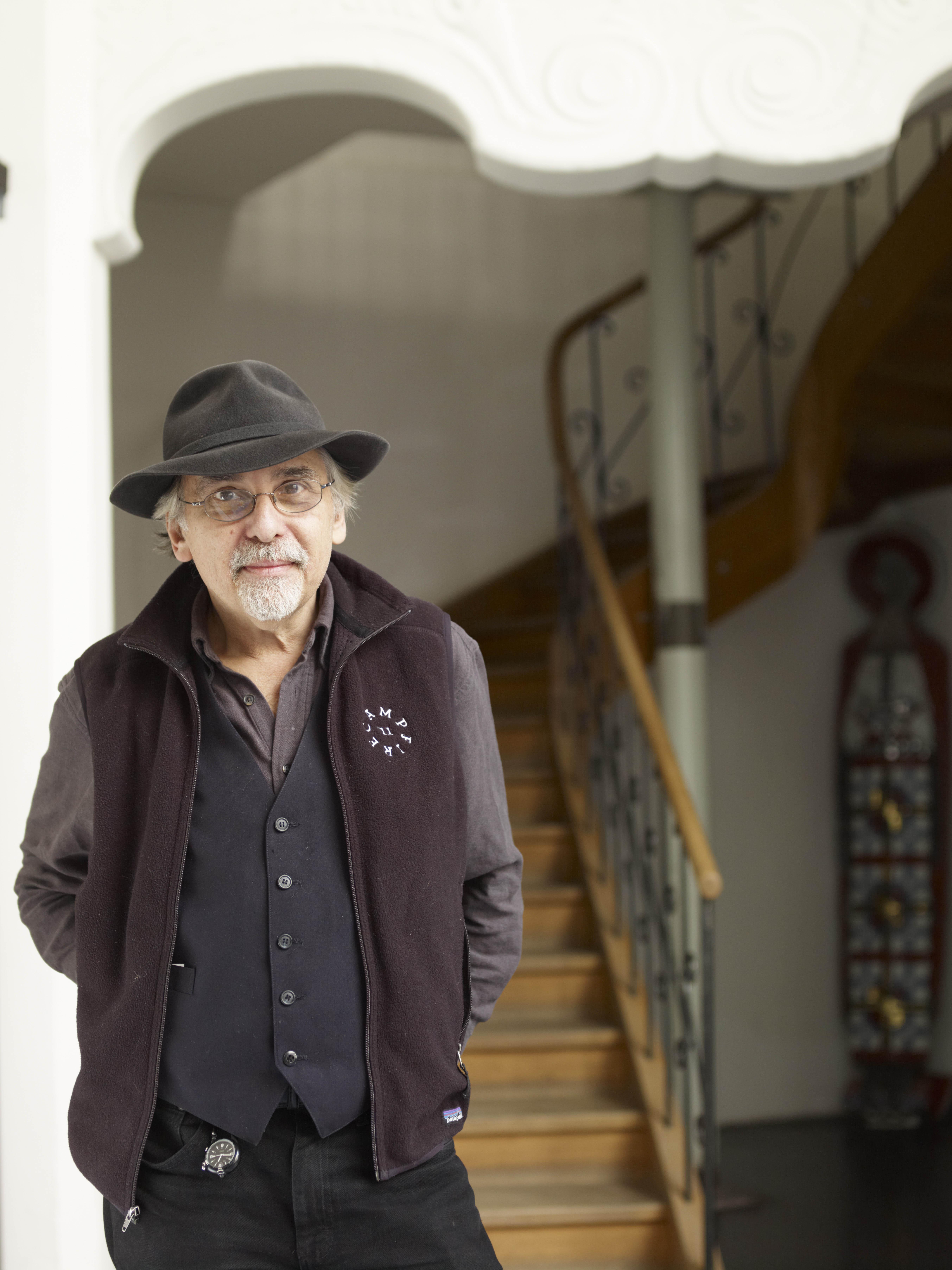 An older man with a white goatee and glasses is dressed in black and brown and wears a felt hat.