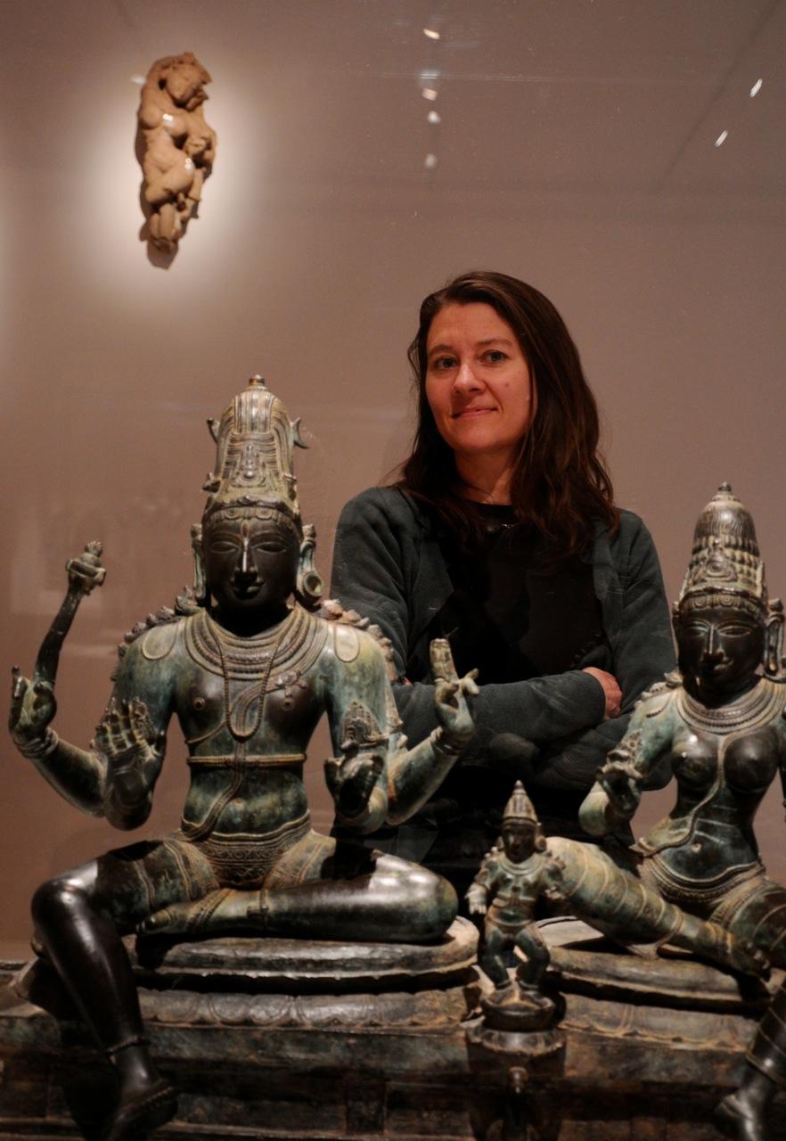 A dark haired, light skinned woman, with arms folded, stands behind two East Asian sculptures of seated figures.
