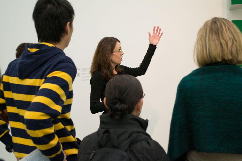 A female tour guide gestures to her left in front of several onlookers.