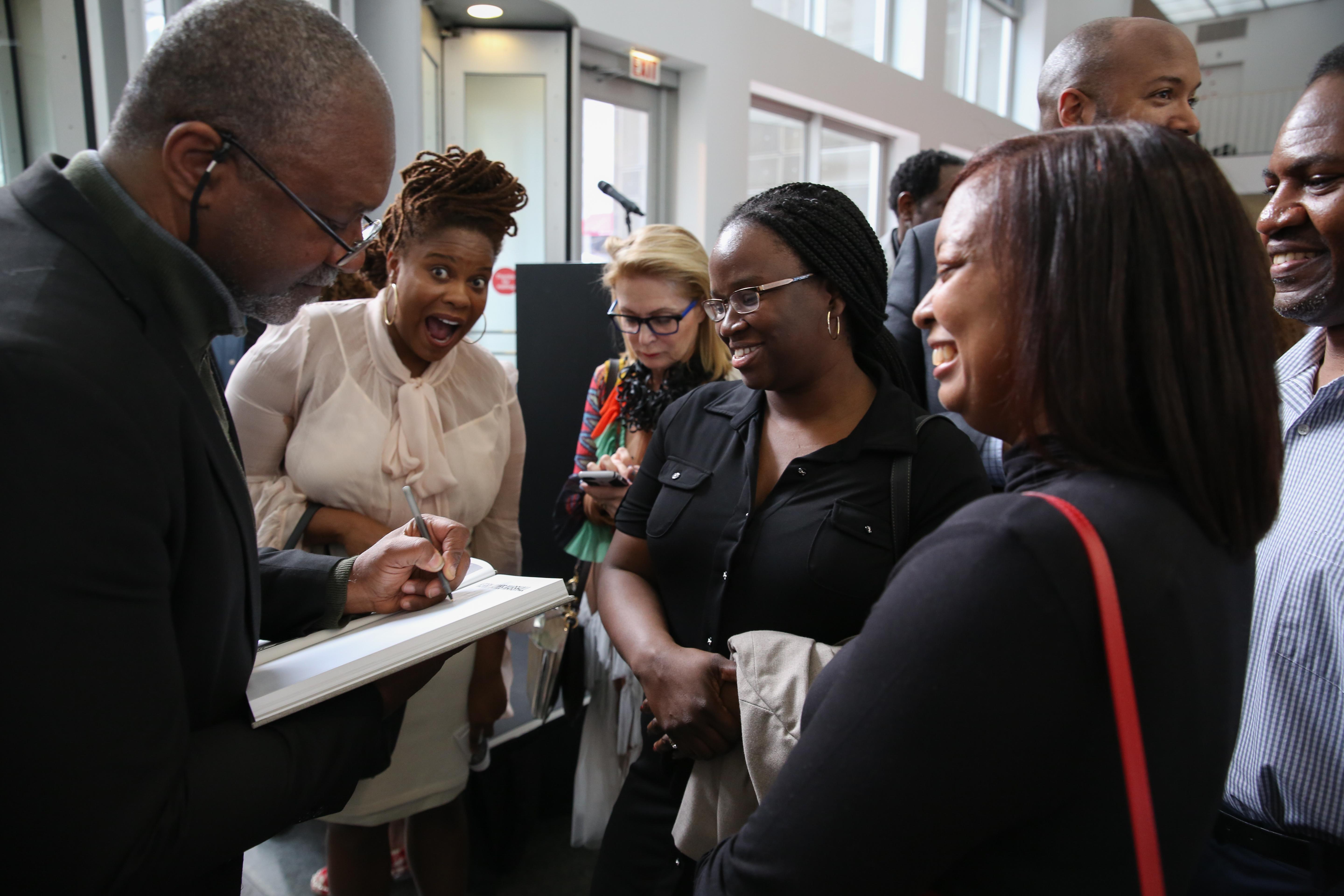 Smiling people gather around Kerry James Marshall as he signs a book. A woman in the center mugs exuberantly for the camera.