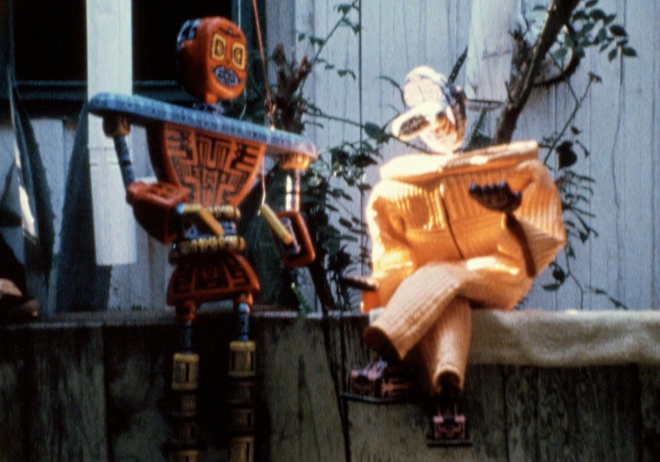 Two orange robots sit outdoors facing each other as if in conversation.