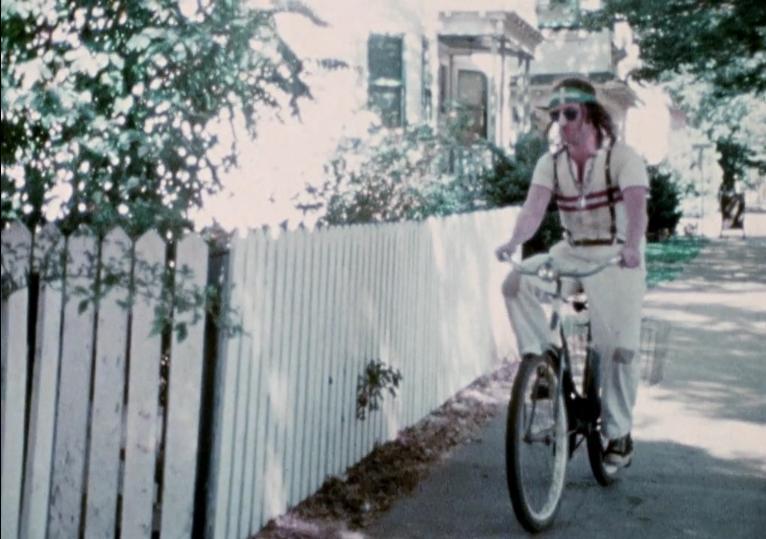 A grainy video image of a person riding a bike in front of a white picket fence.