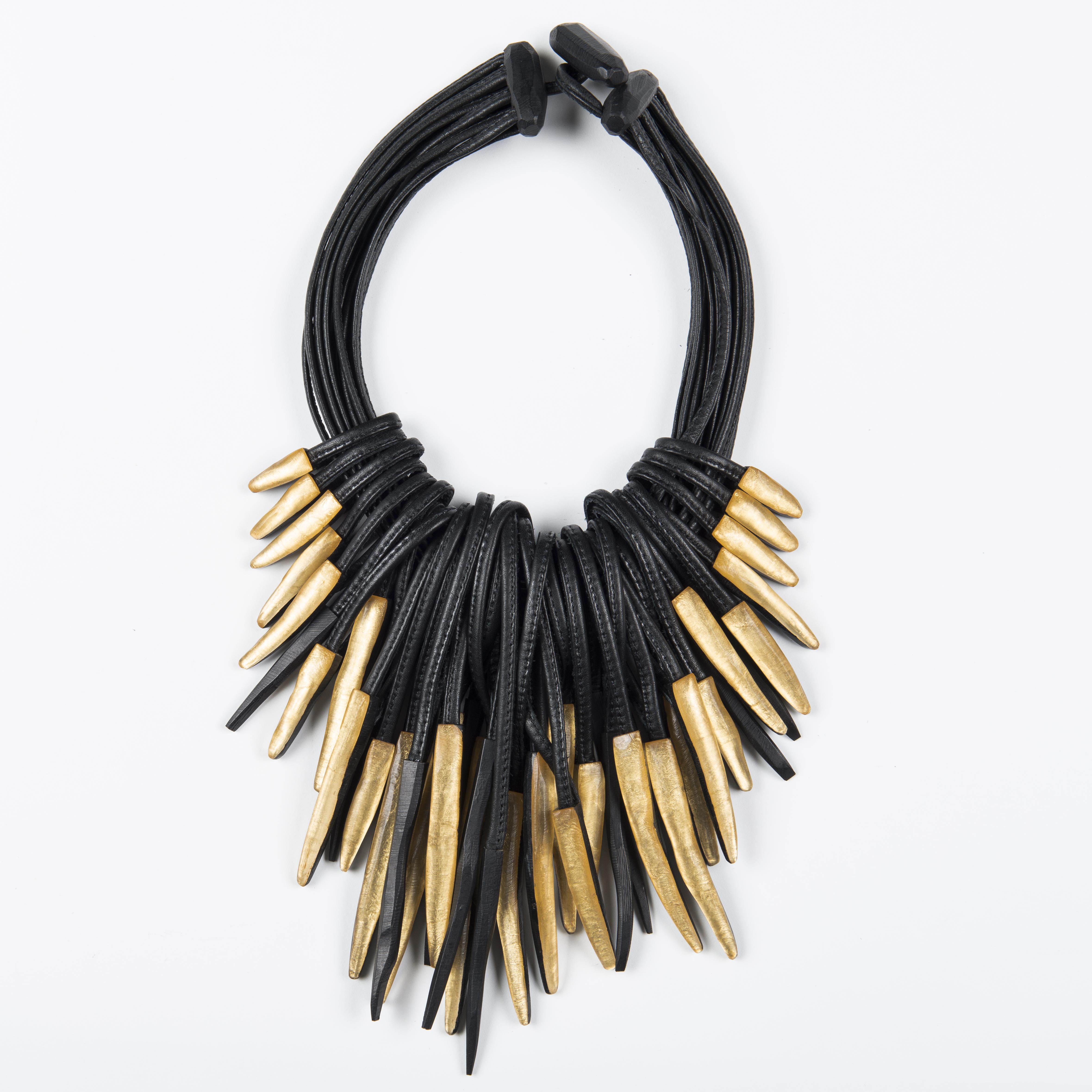 A necklace made from thick black leather cords features many gold dart-like pendants and fastens with a toggle at the top.
