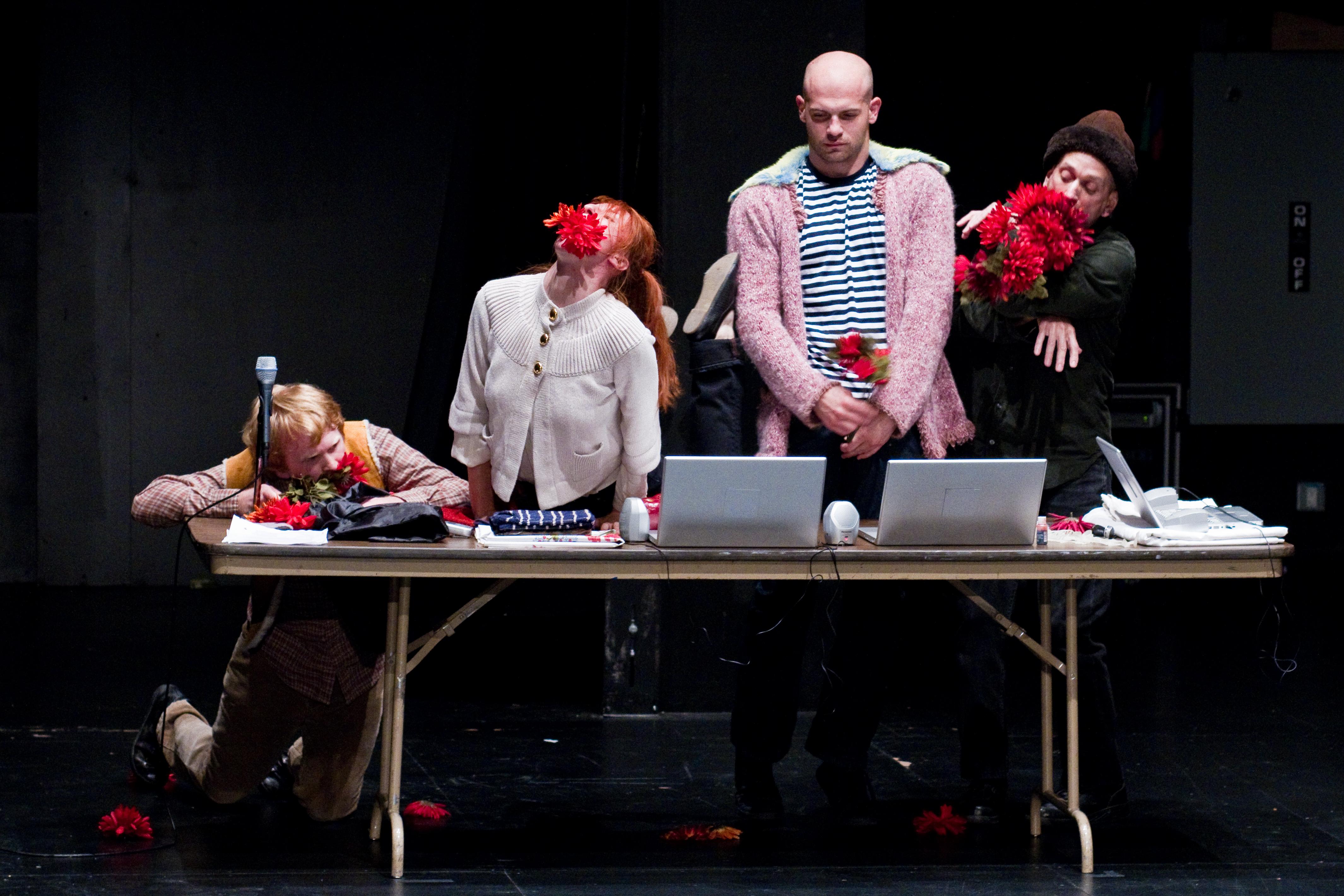 Three men and one woman stand or rest behind a rectangular, collapsible table with three laptops and various objects.Two people hold red flowers in their hands and arms; two have red flowers in their mouths.
