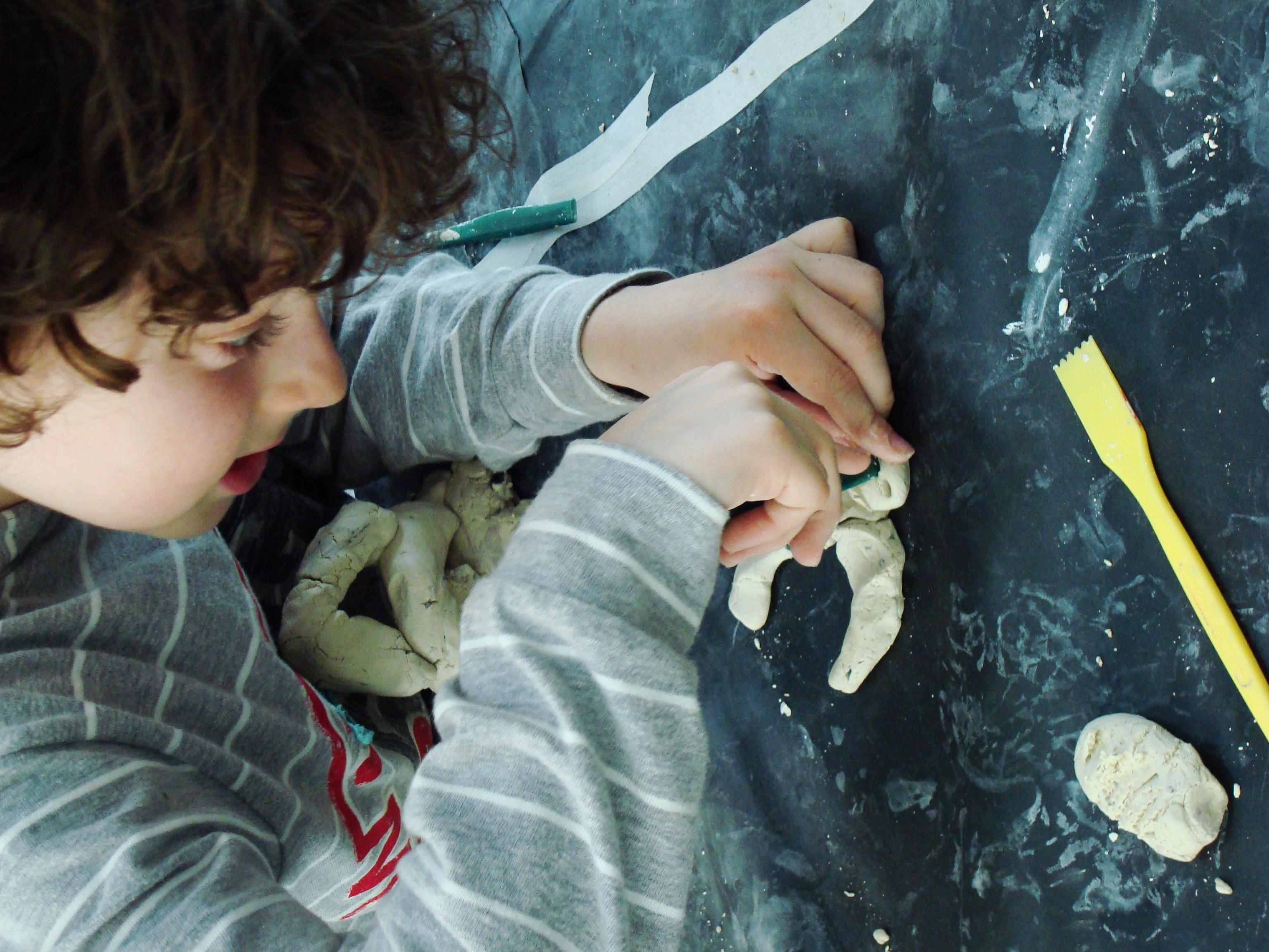 A pale-skinned child with short, dark hair is bent over, creating art with clay and tools.