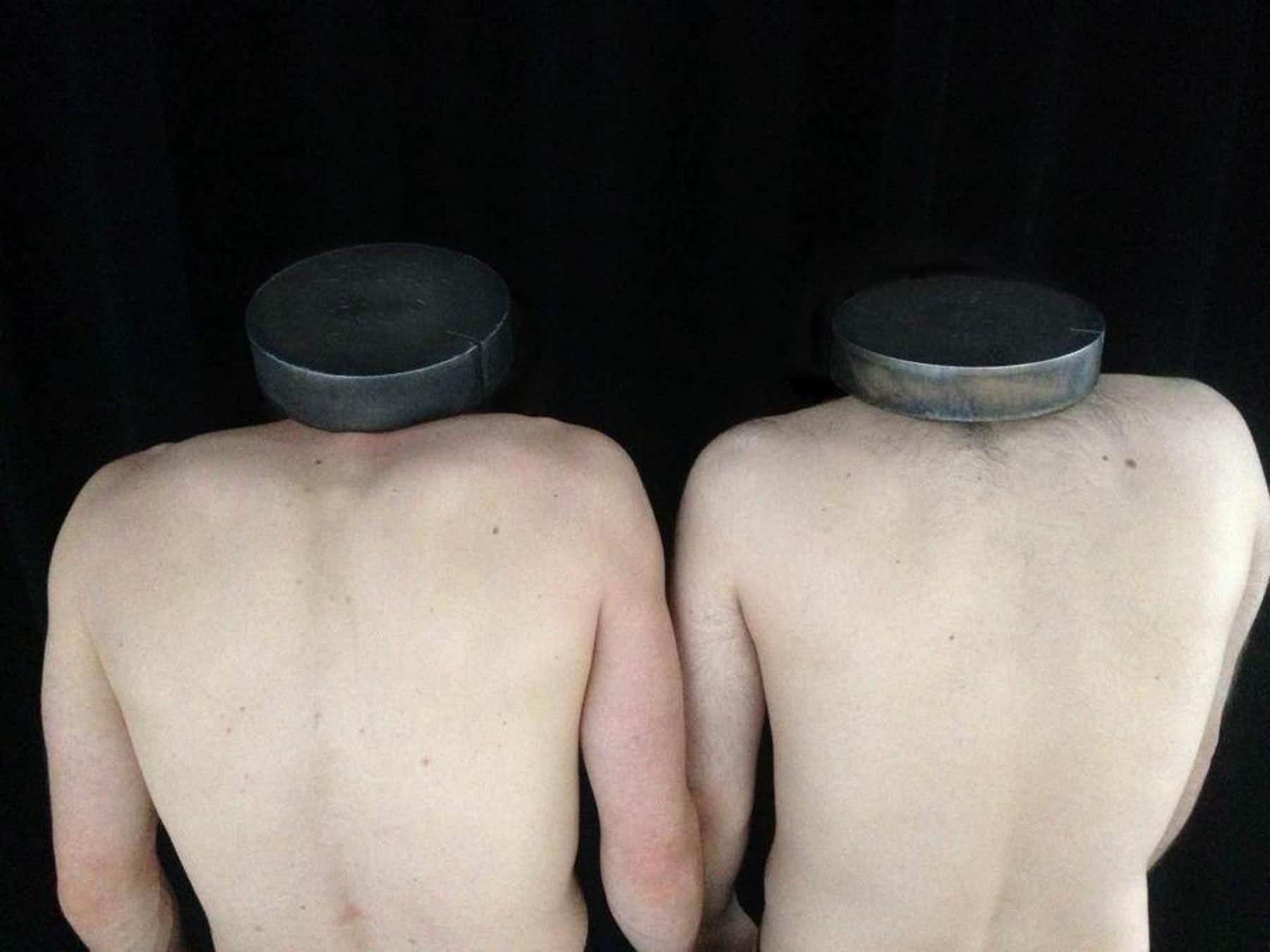 Photograph of two nude headless male torsos seen from behind against a black background. Round metal discs rest on their shoulder in place of heads.