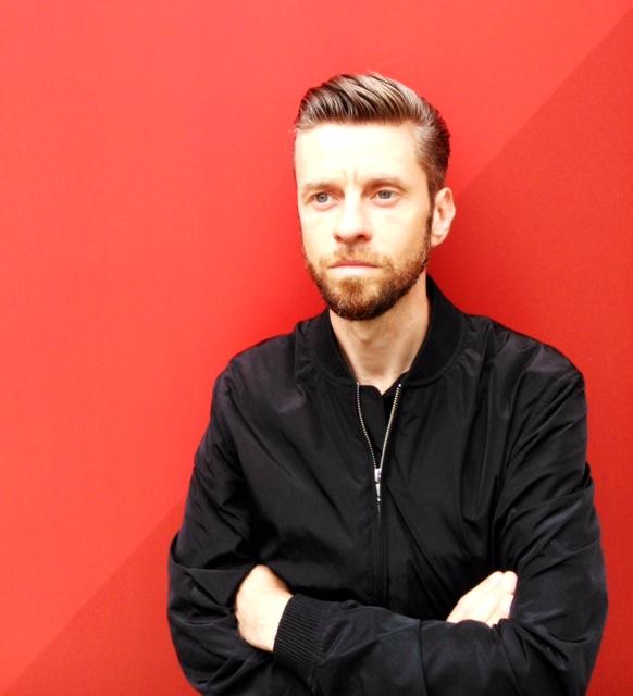 A light-skinned man dressed in all black with slicked-back hair and a beard poses with arms crossed in front of a red background.