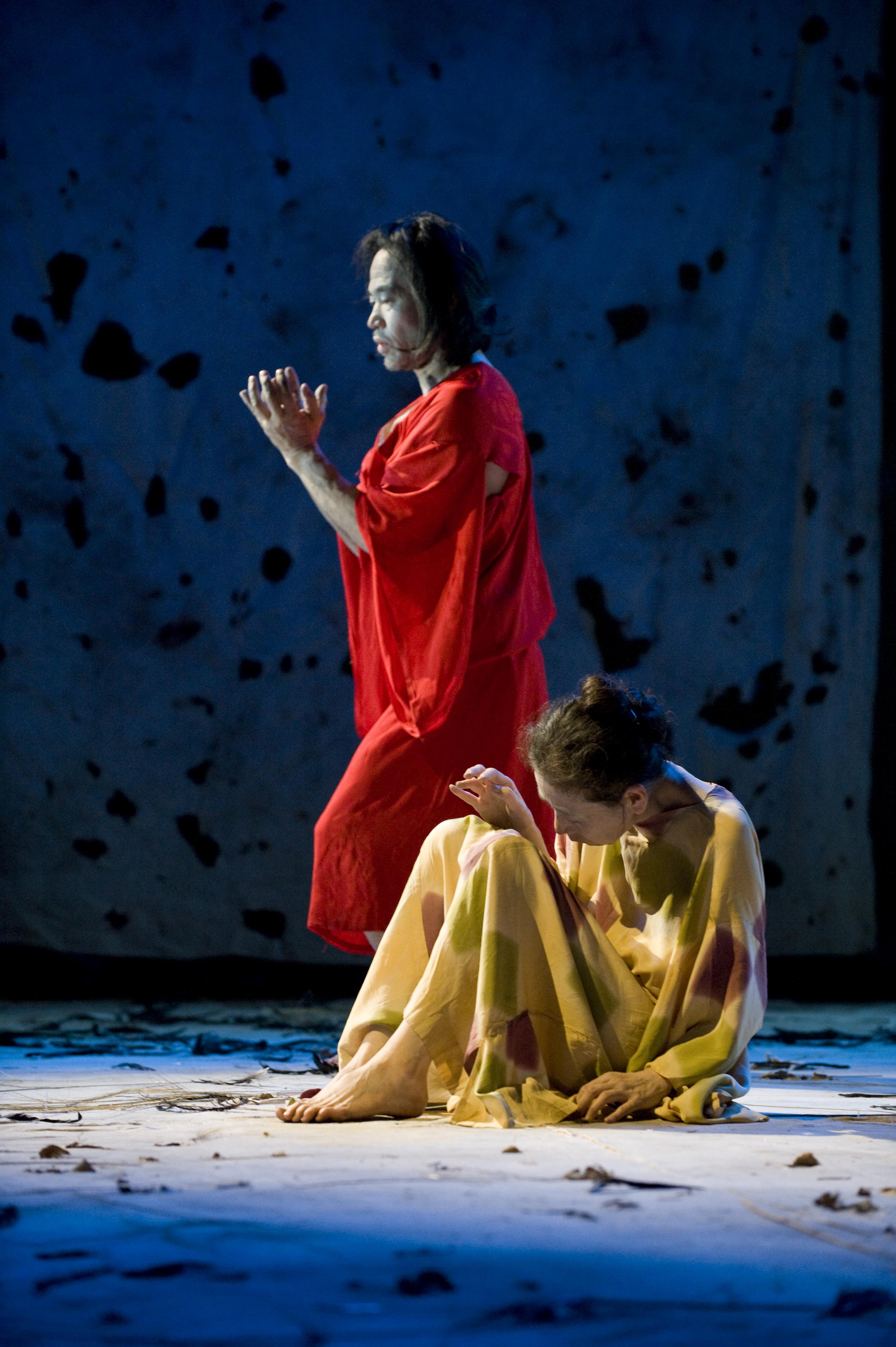 A figure clad in a loose red robe stands behind a seated figure in a loose yellow robe. The stage and backdrop behind them are blue, spotted randomly with dark stains.