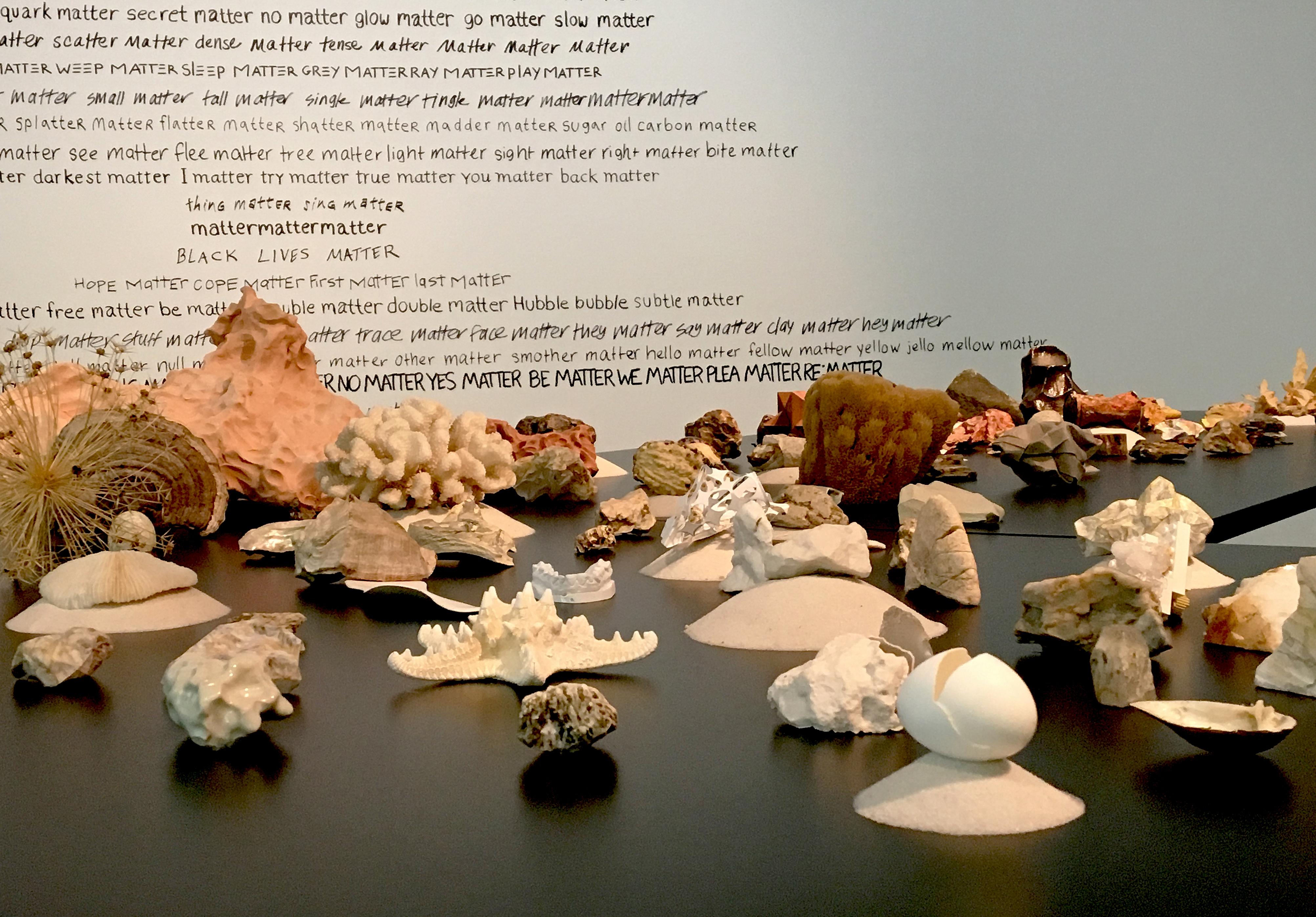 A table with rocks, minerals, and other substances sits in front of a wall covered with black text.