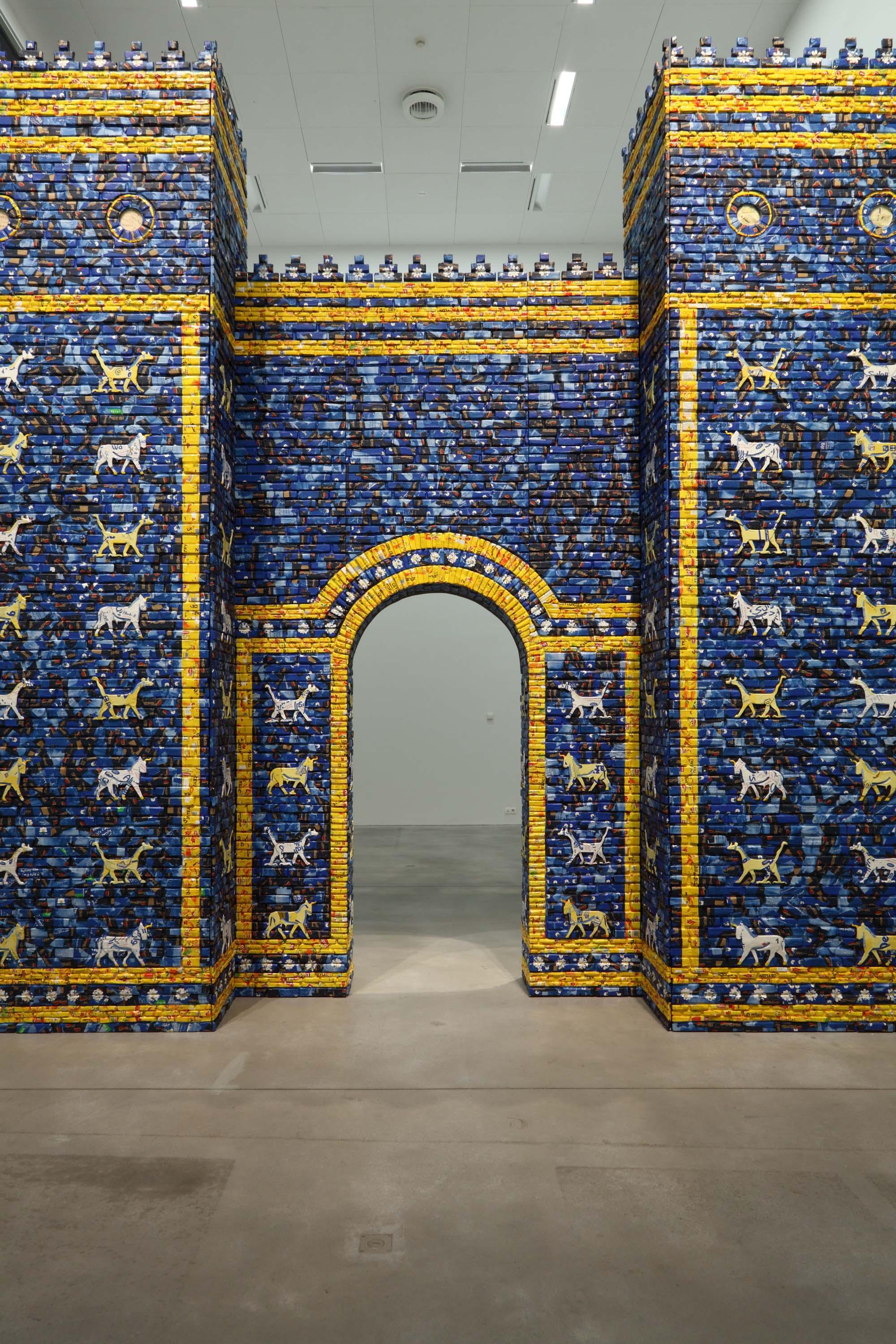 An architectural arched gateway is shown, installed within a modern gallery space. The arch is made from bricks in a range of different blues with golden yellow accents and decorated with stylized animals.