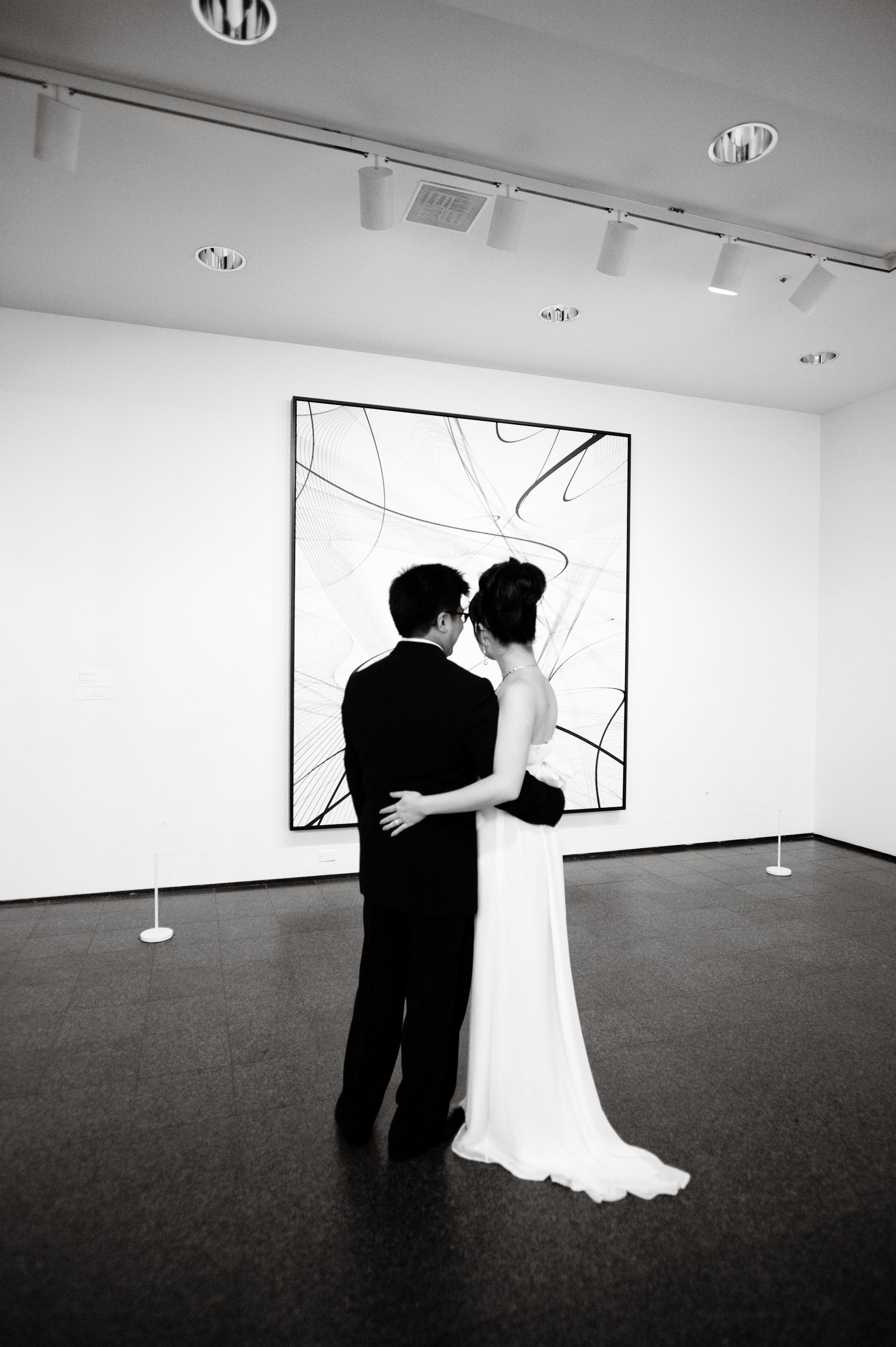 A bride and groom embrace in front of an artwork, with their backs to the camera.