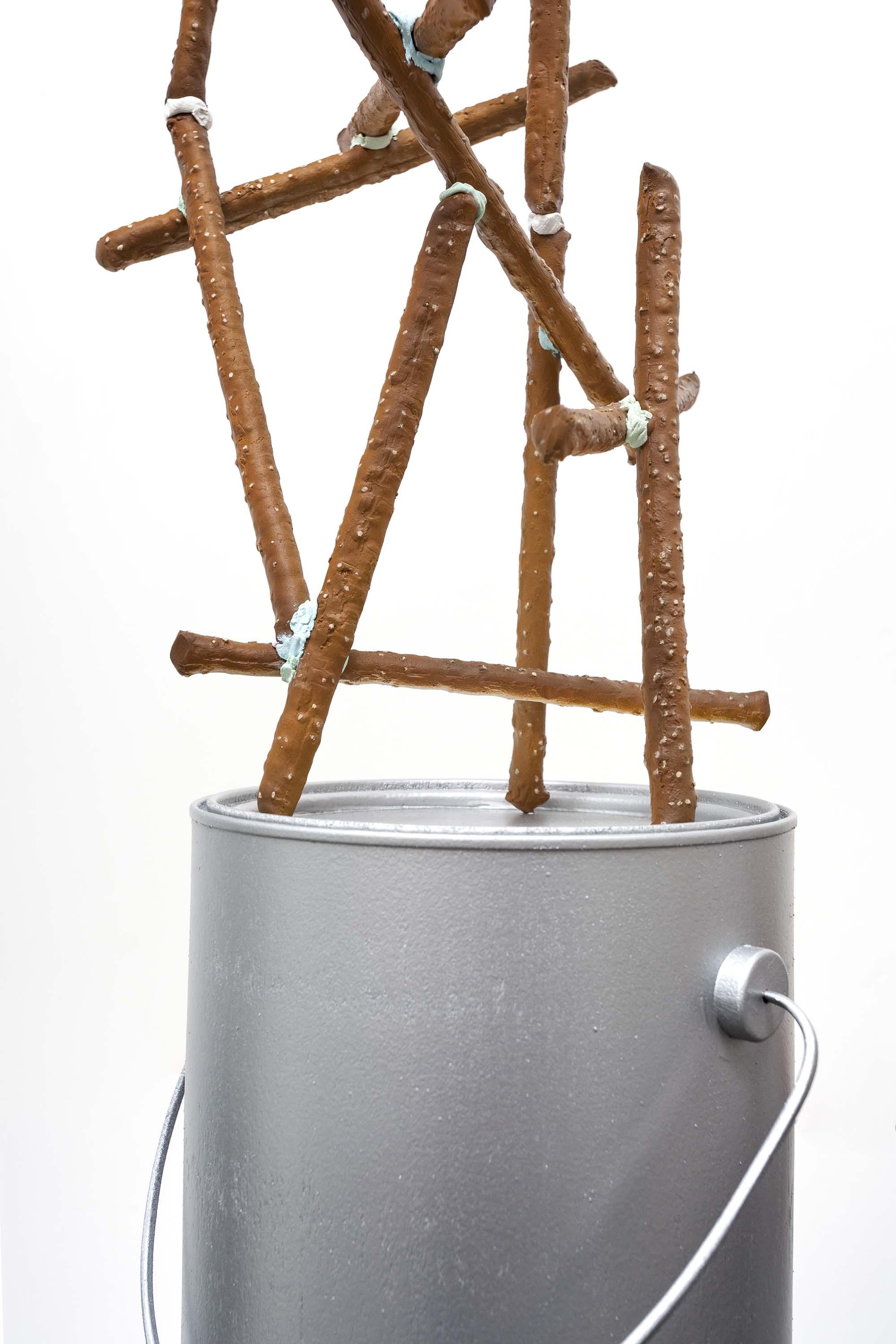 Brown pretzel sticks with specks of white are glued together in a stacked formation, and are balanced atop a silver paint can.