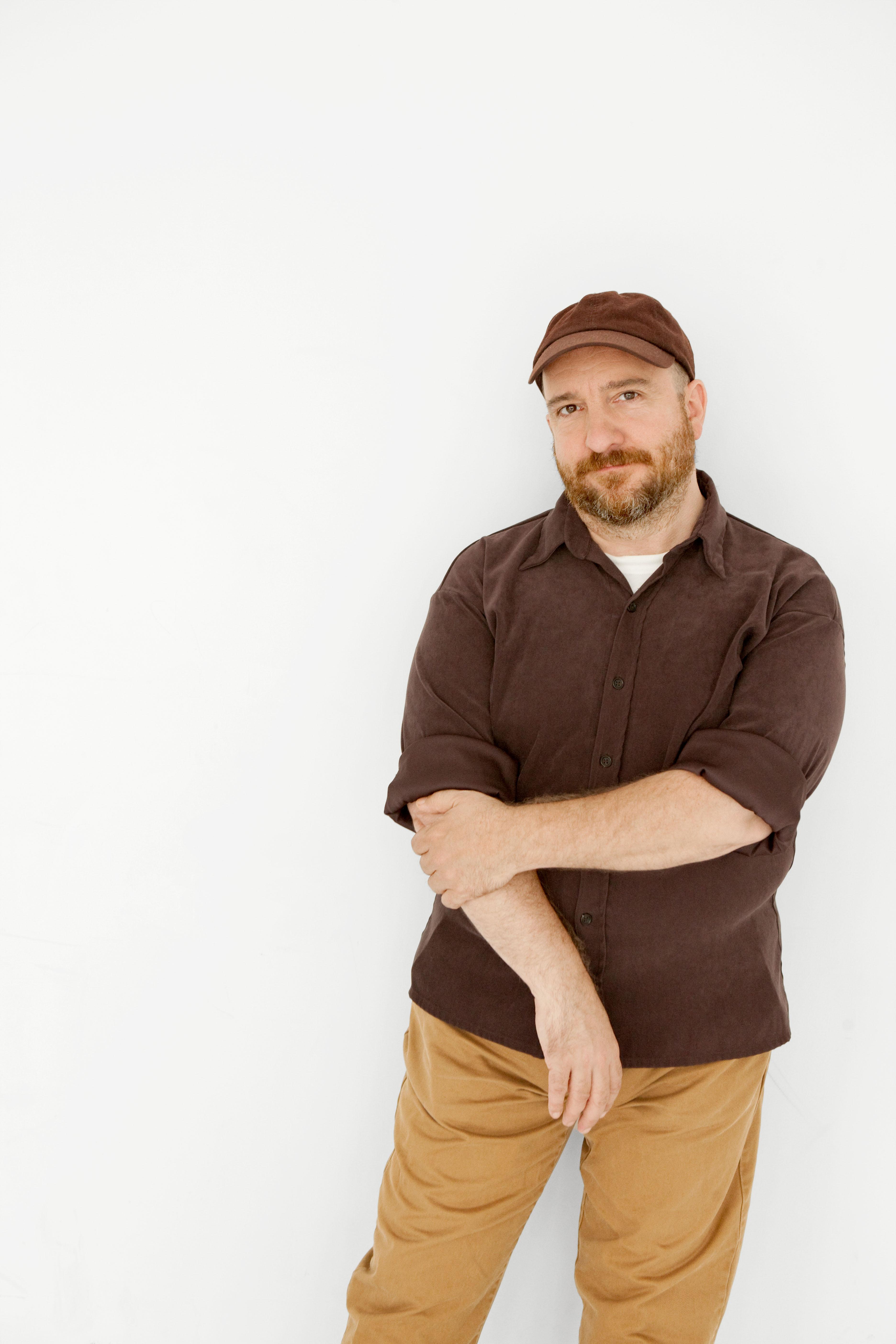 A light-skinned man in casual dress poses standing. He looks directly at the camera with a neutral expression.