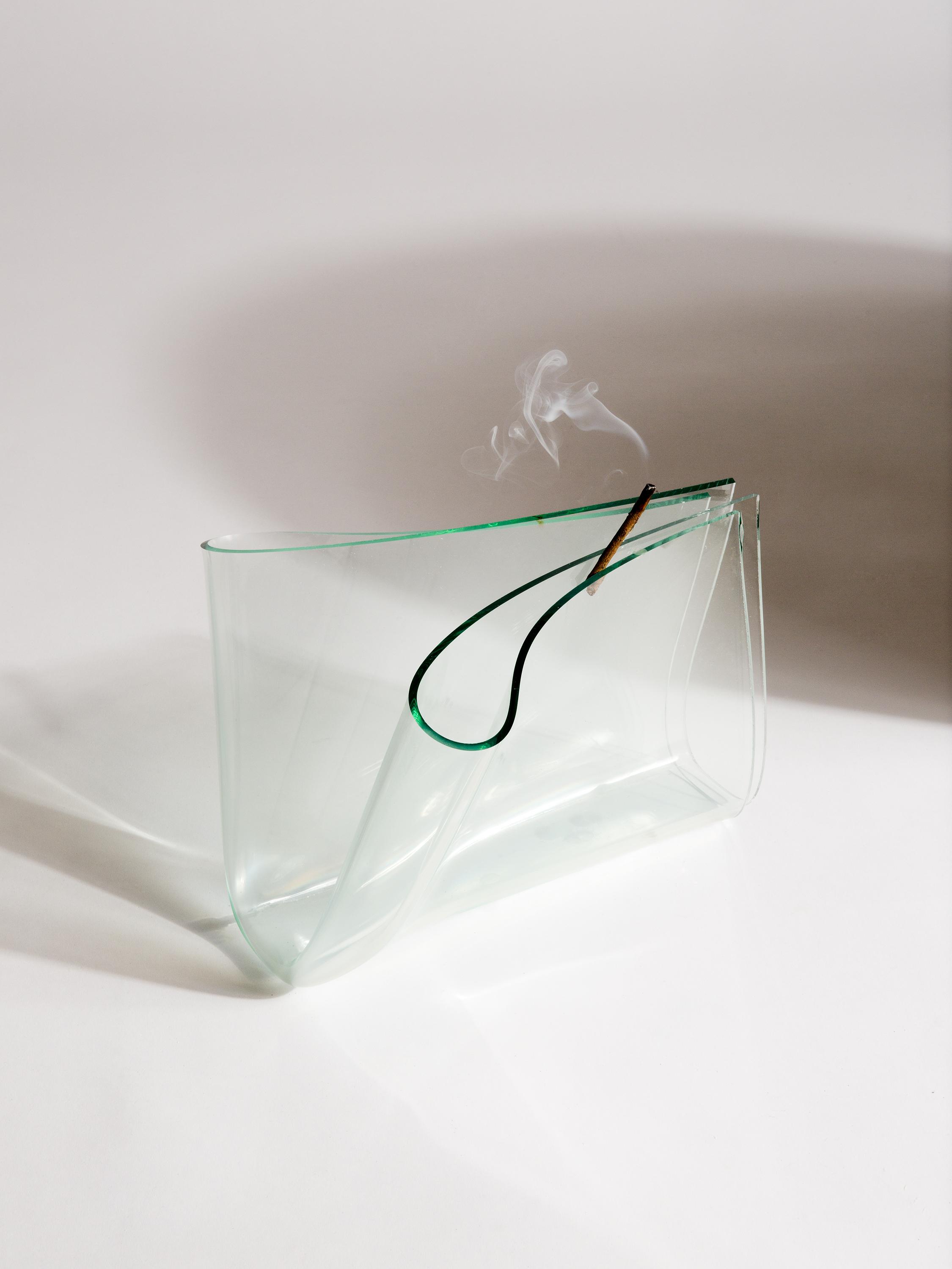 A piece of glass is folded into quarters with green edges facing up, resting on a white surface. A smoking stick is wedged into the top fold. There is a dark, round shadow behind the sculpture on the right.