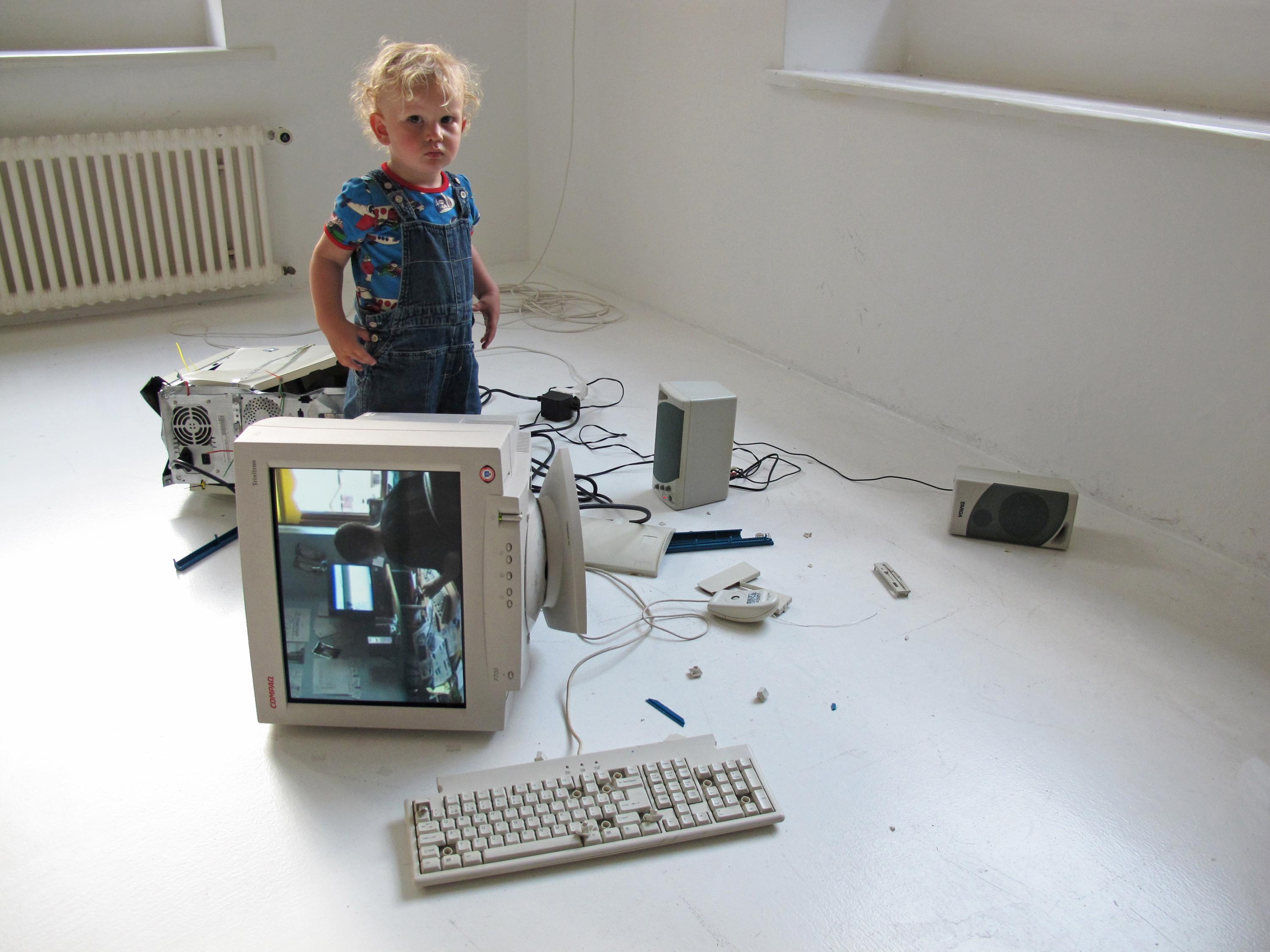 A blond toddler stands in a white room amid a mess of upturned, damaged computer equipment.