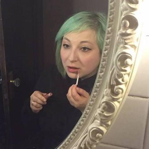 A young, light-skinned woman with with pastel-green hair applies lip gloss while looking in a mirror with an ornate white frame.