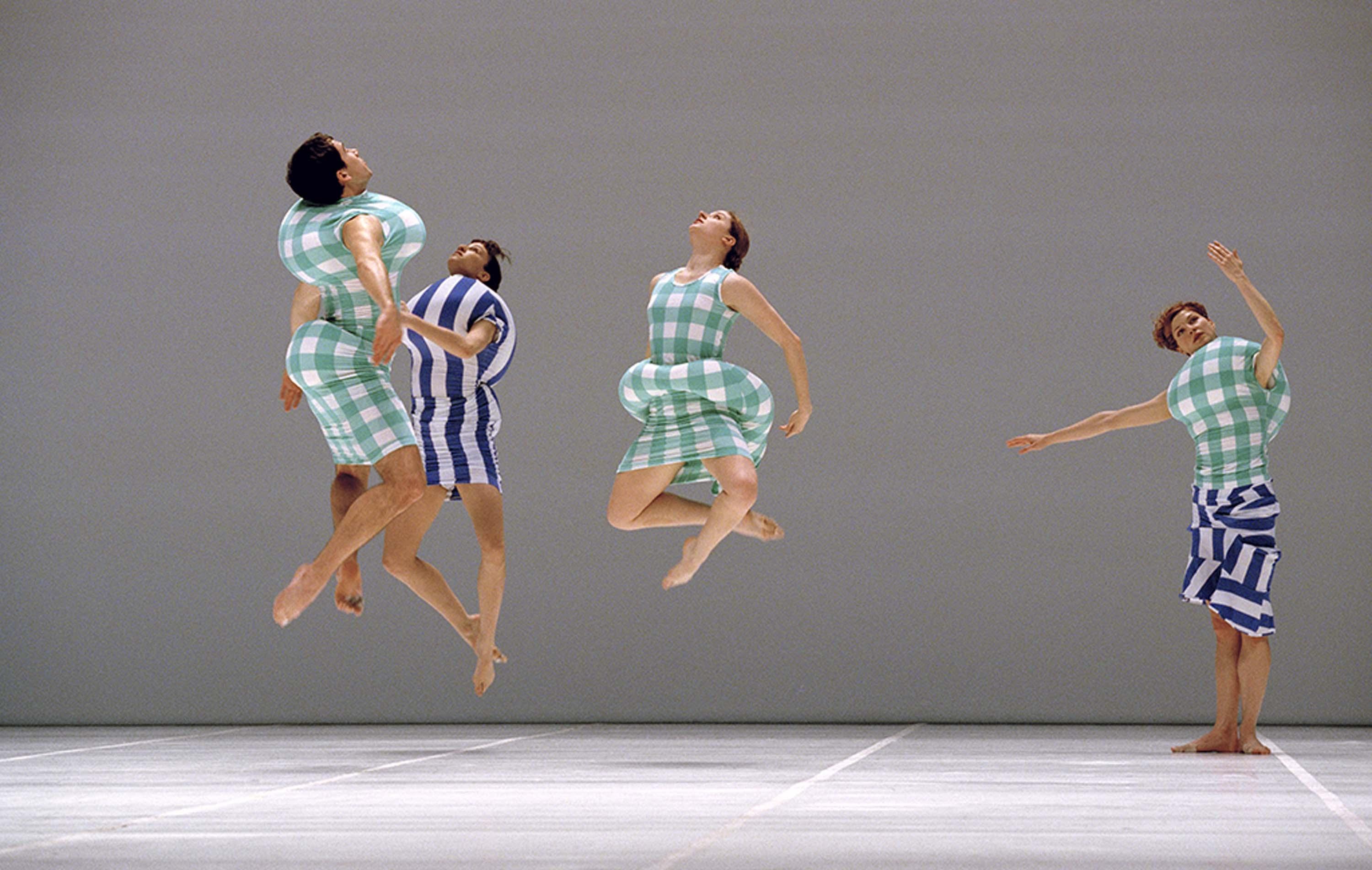 Three dancers leap in the air while looking up, while a fourth dancer, frozen in a robotic gesticulation, stands to the right against a grey backdrop. They wear padded costumes in bright blue-and-white stripes or green-and-white checks.