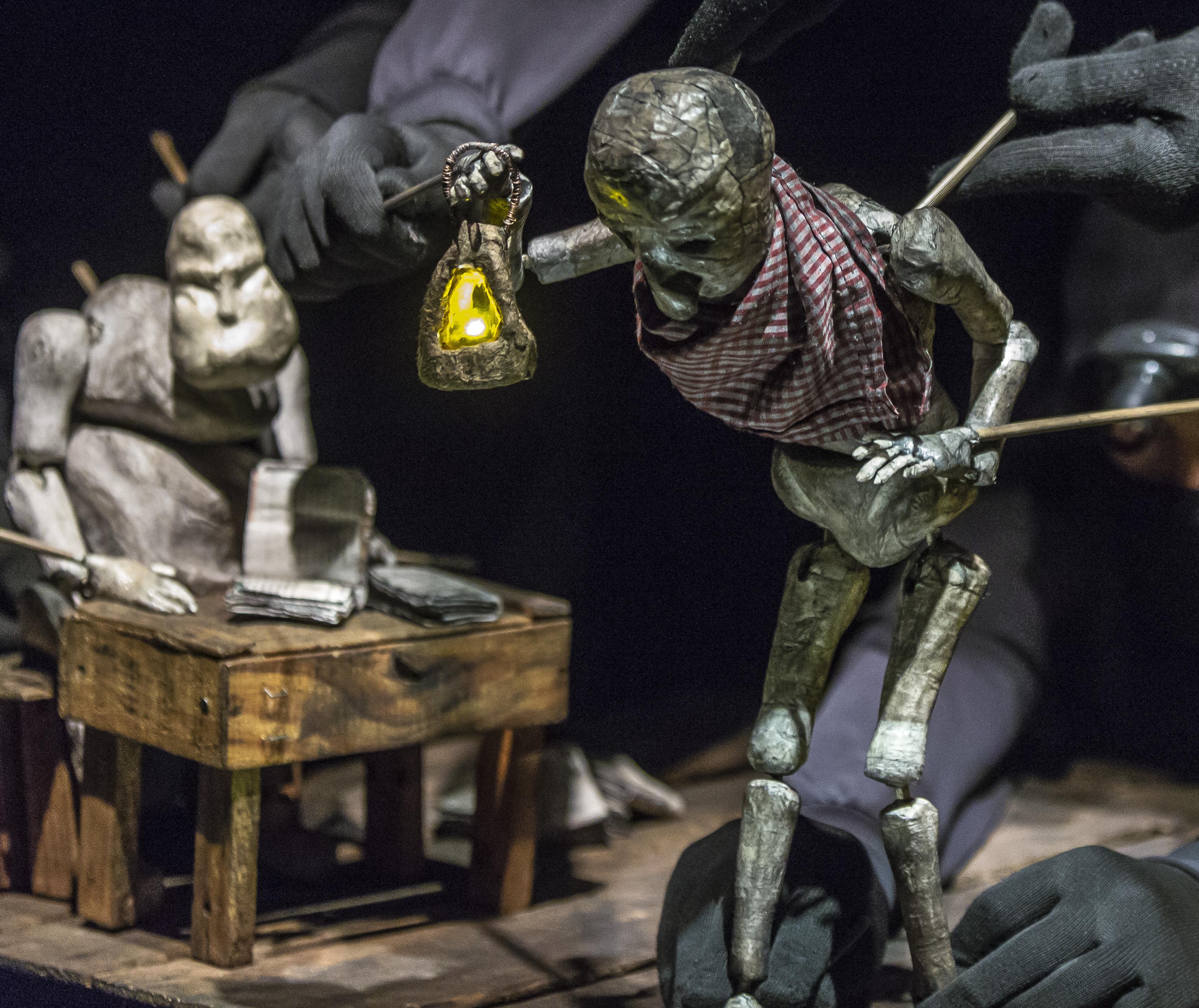 A small coal smudged, paper puppet holds a lantern, while another puppet sits at a table with a book. The gloved hands of the puppeteers manipulate the puppet with thin sticks.