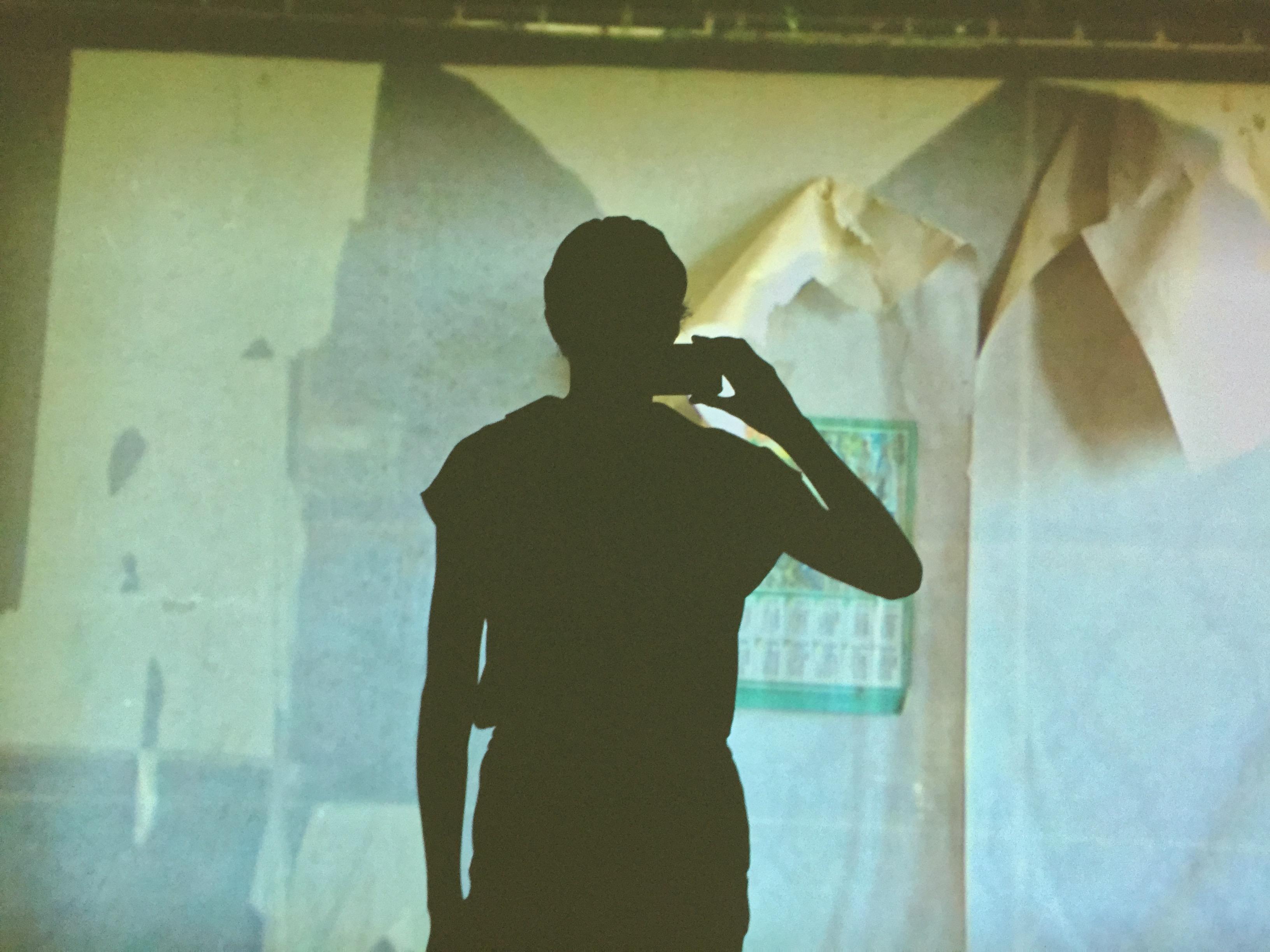 A silhouette of a person photographing a video projection on a wall. The video projection appears to be a room of peeling wallpaper.