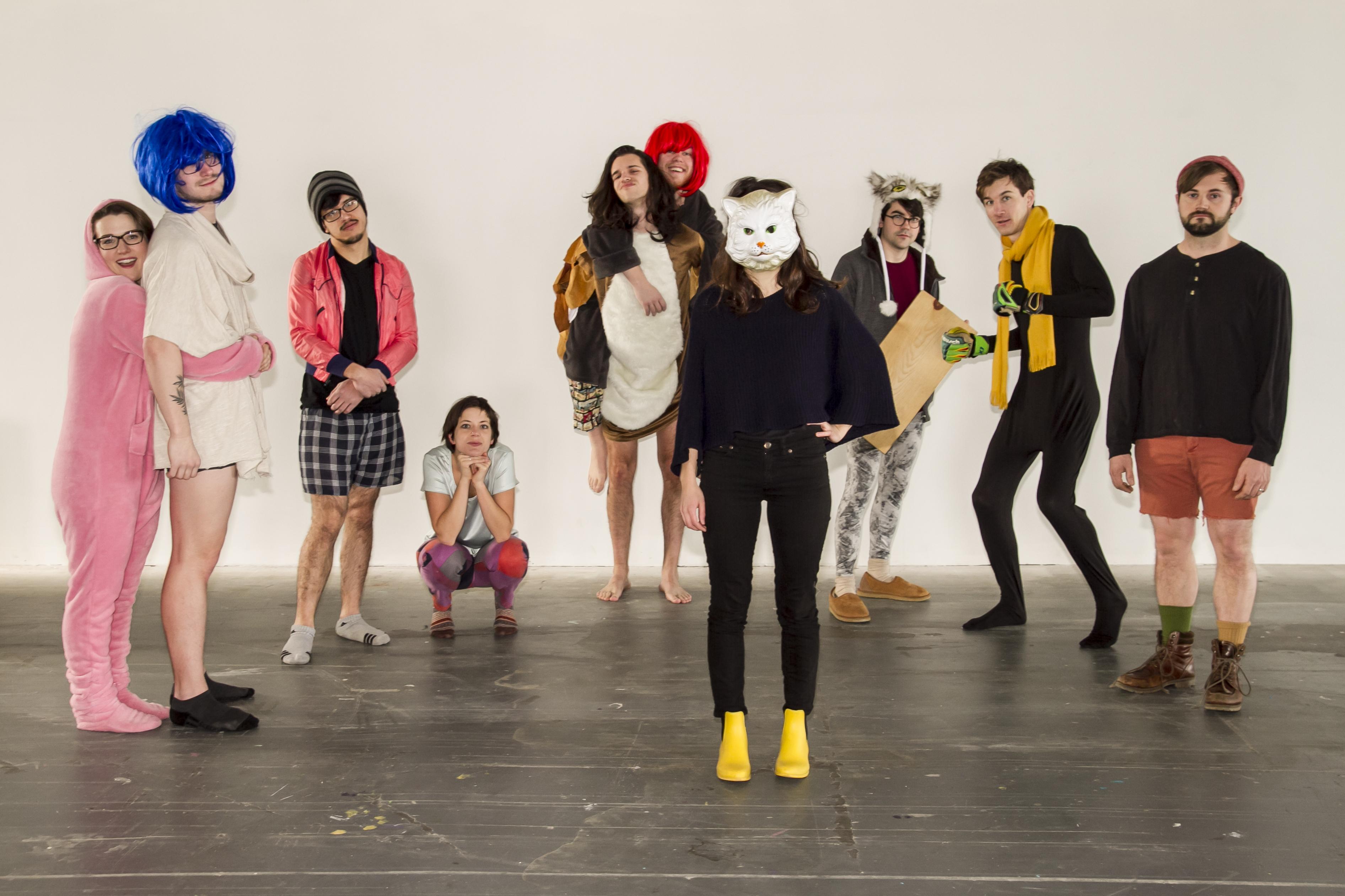 A group of young people pose playfully in an empty room standing in an semi-circle behind one person wearing a cat mask, black shirt and pants, and bright yellow shoes. Some wear brightly colored wigs and colorful clothing.