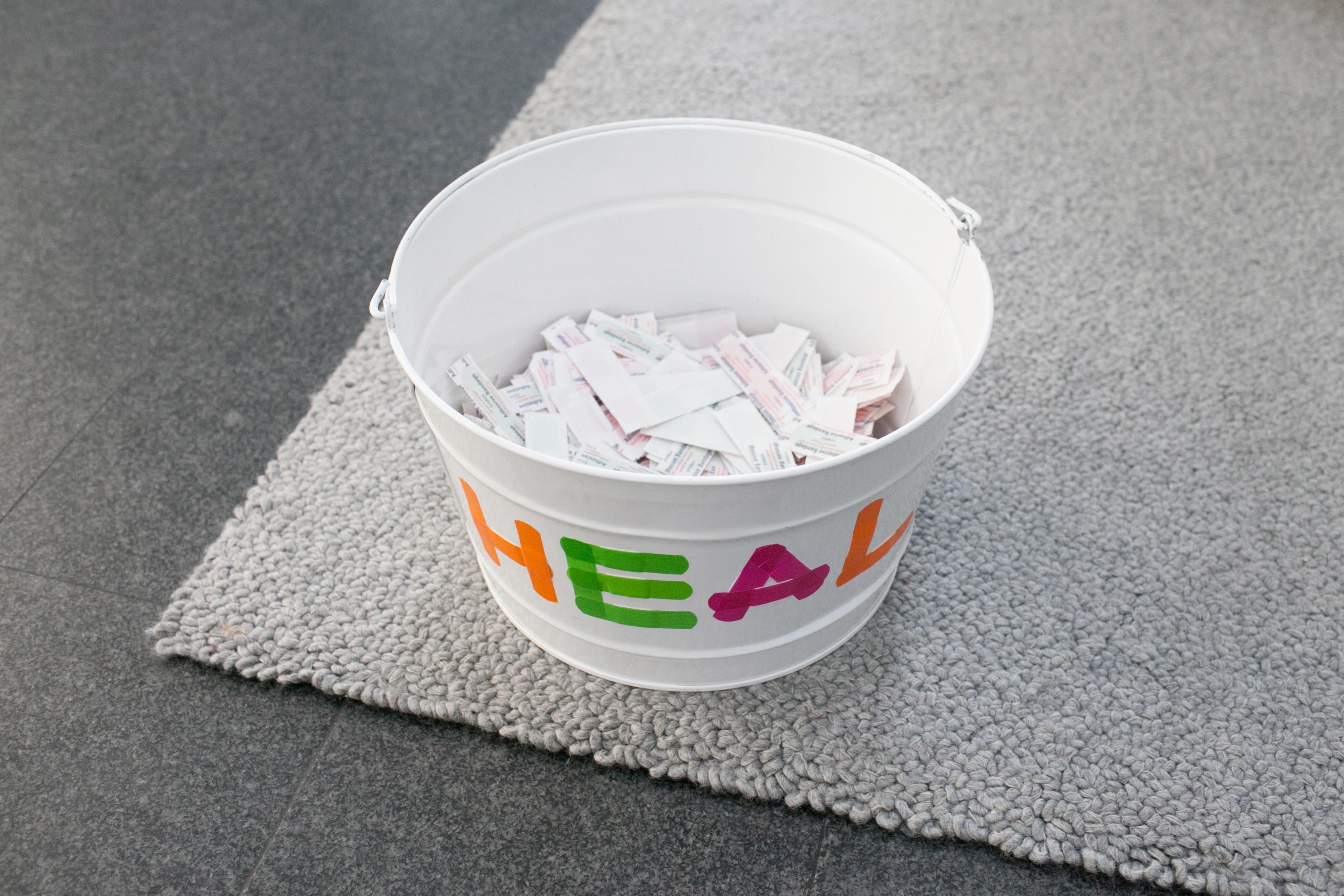 A white pail labeled "HEAL" in bright colors sits on the corner of a gray rug, which in turn rests on a gray granite floor. The pail is half-way filled with slips of paper.