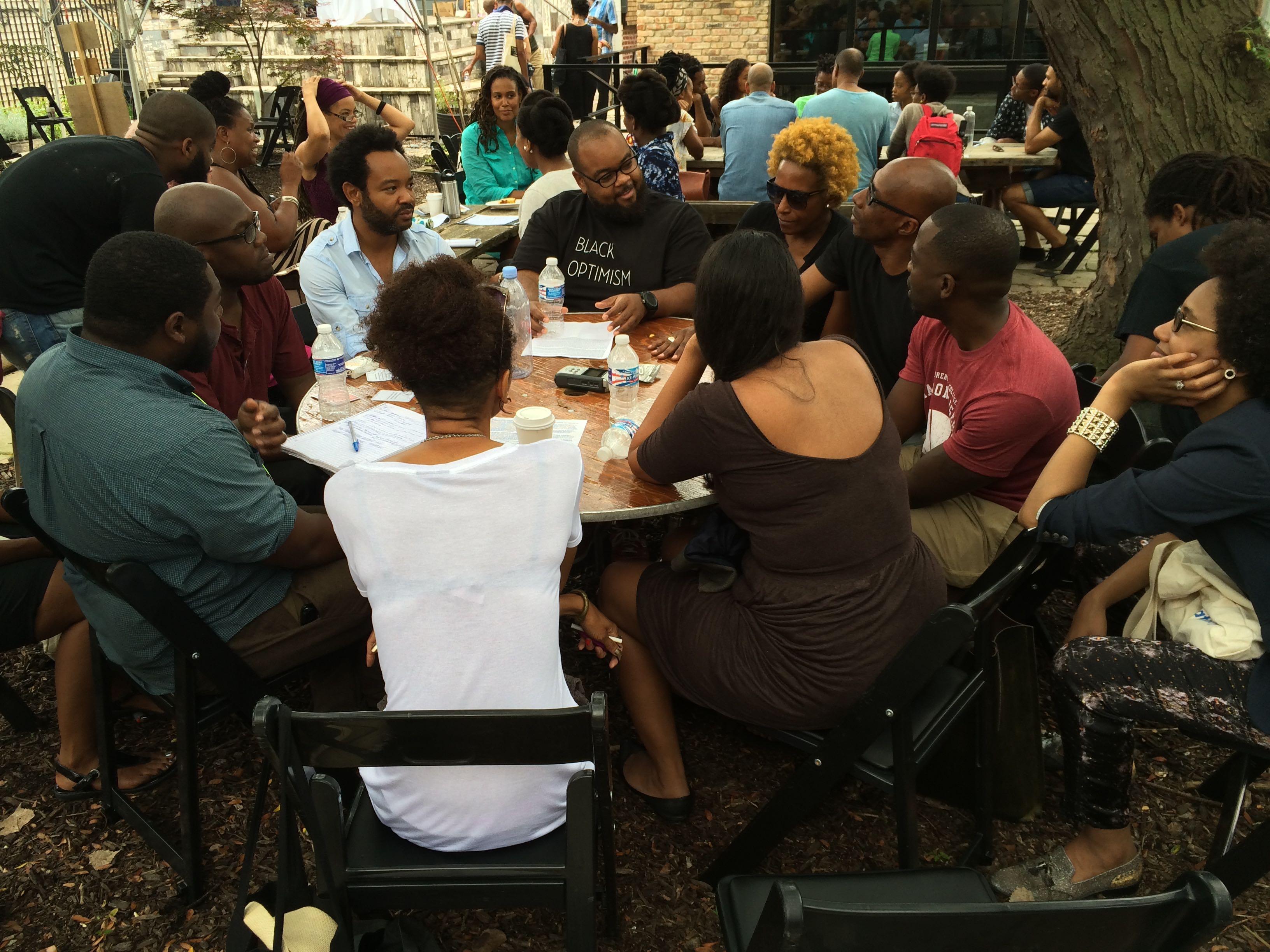 A group of dark-skinned people sit talking around a table outdoors, with other tables of people visible in the background.