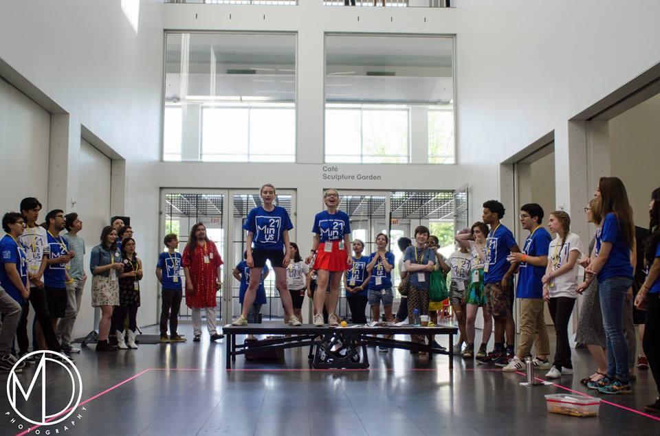 Two teens stand on a platform in the MCA's atrium surrounded by a crowd of young people.