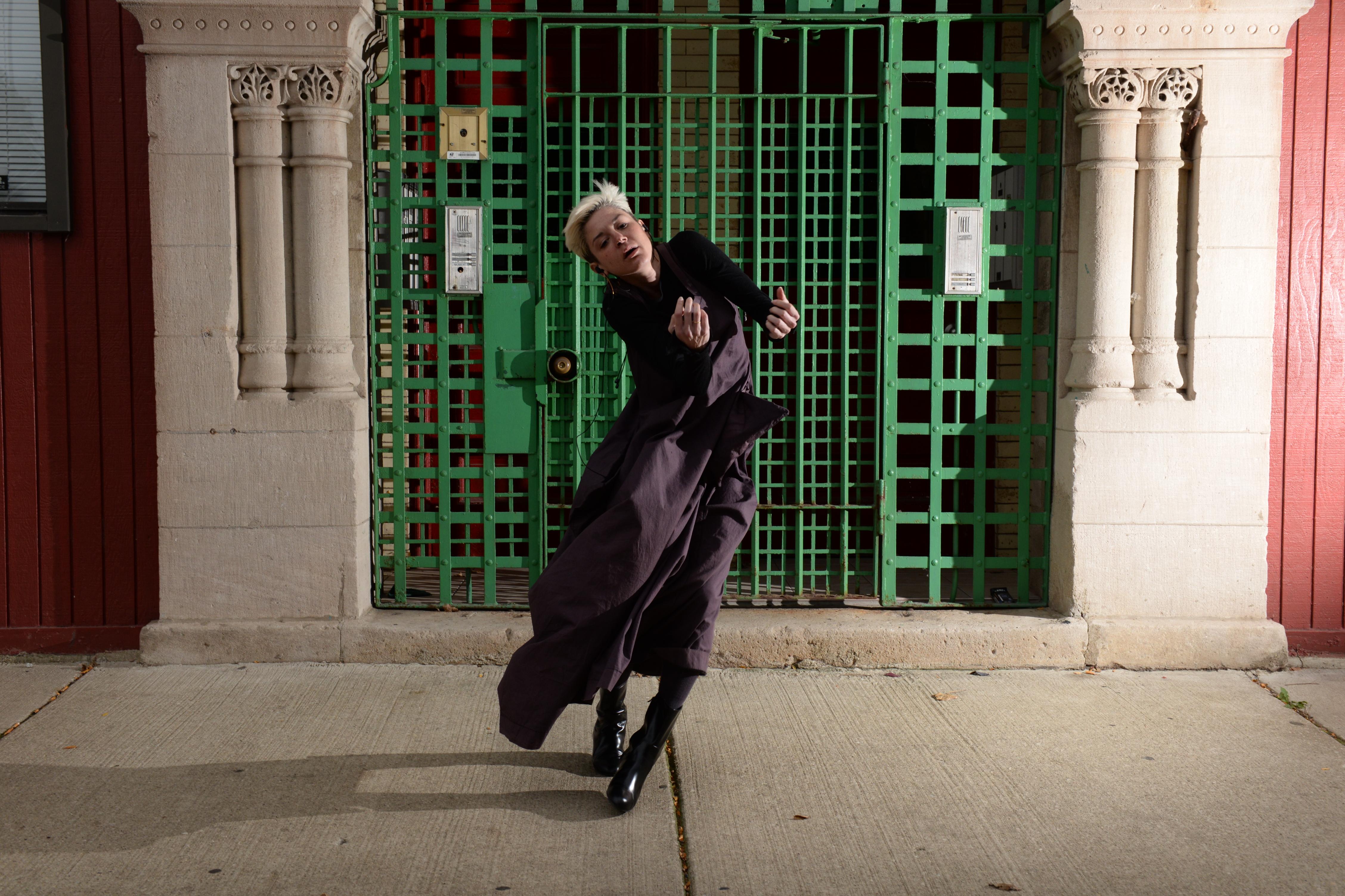 A person wearing a long purple jacket and black boots strikes a dance pose on a sidewalk in front of a stone building with a green gridded gate blocking the entrance.