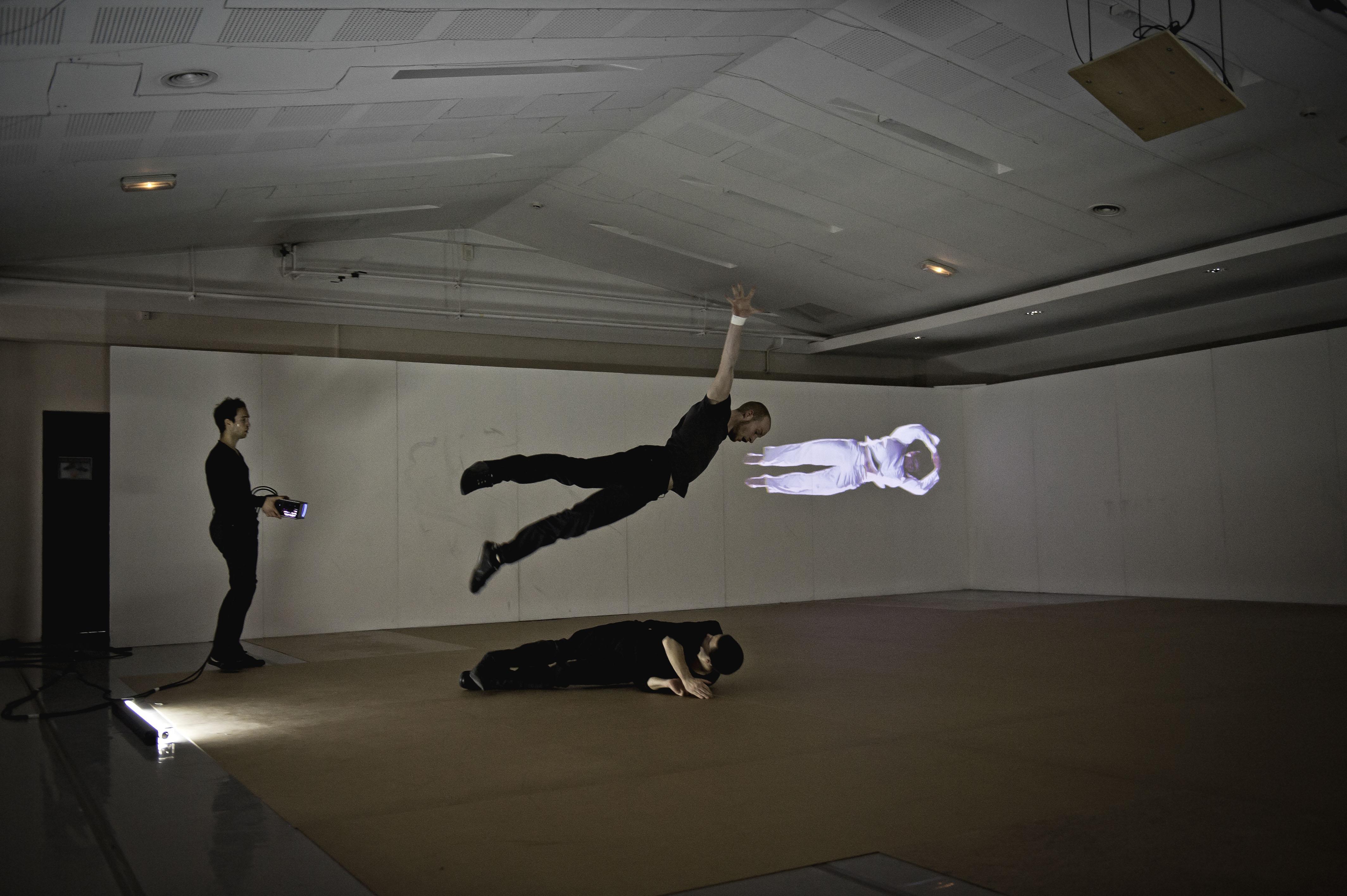 A man is in mid-air, limbs spread, above another figure curled on the floor. A third person holds a projector that casts an image of a person wearing white on the wall.