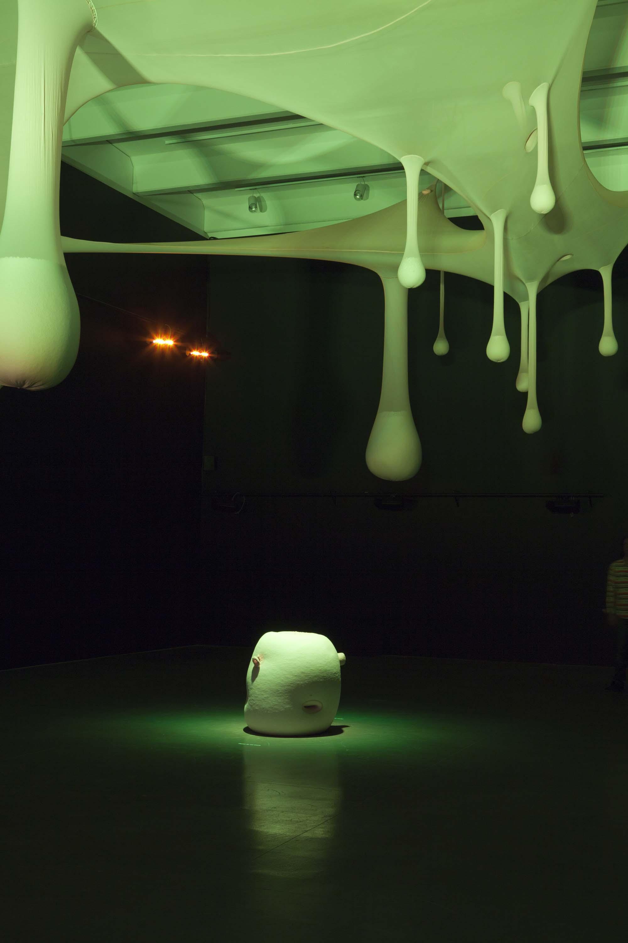 Amorphous fabric shapes hang from the ceiling of a dark gallery above a white sculptural form resting on the floor that has an eerie green glow.