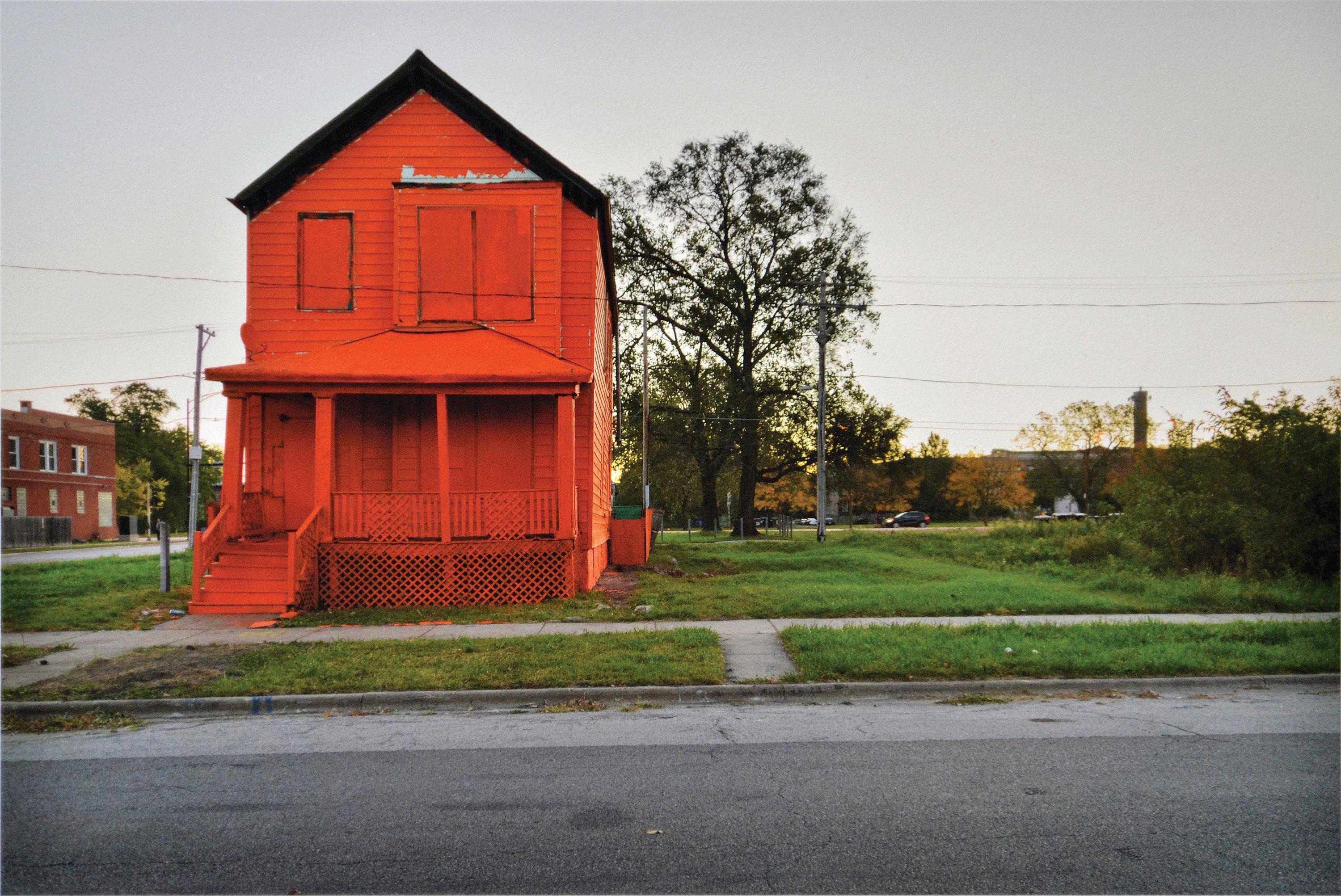 A bright red-orange house is boarded up, standing next to an empty urban lot during sunset.