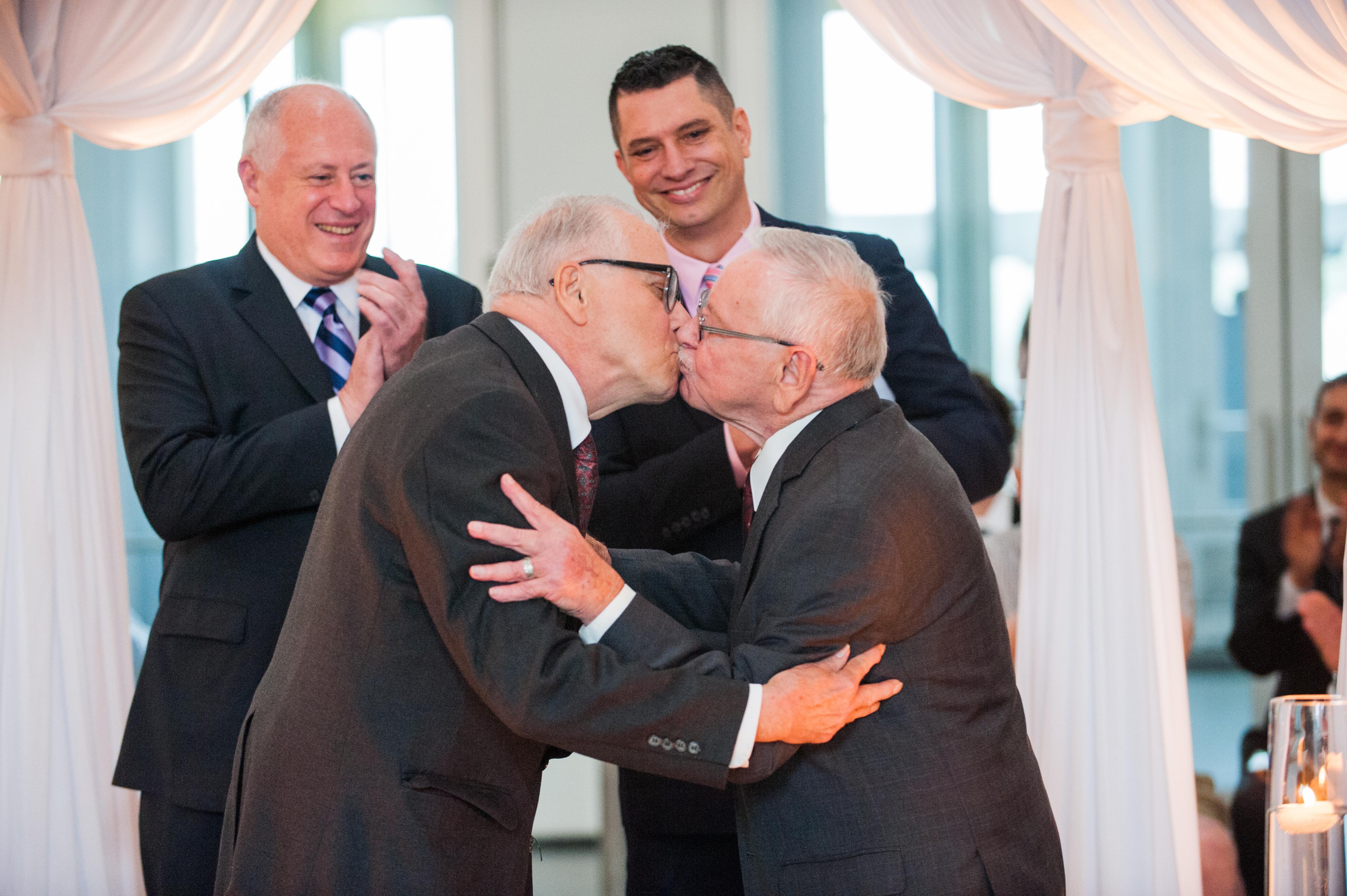 Two elderly men wearing suits embrace in a kiss while two other men look on smiling and clapping.