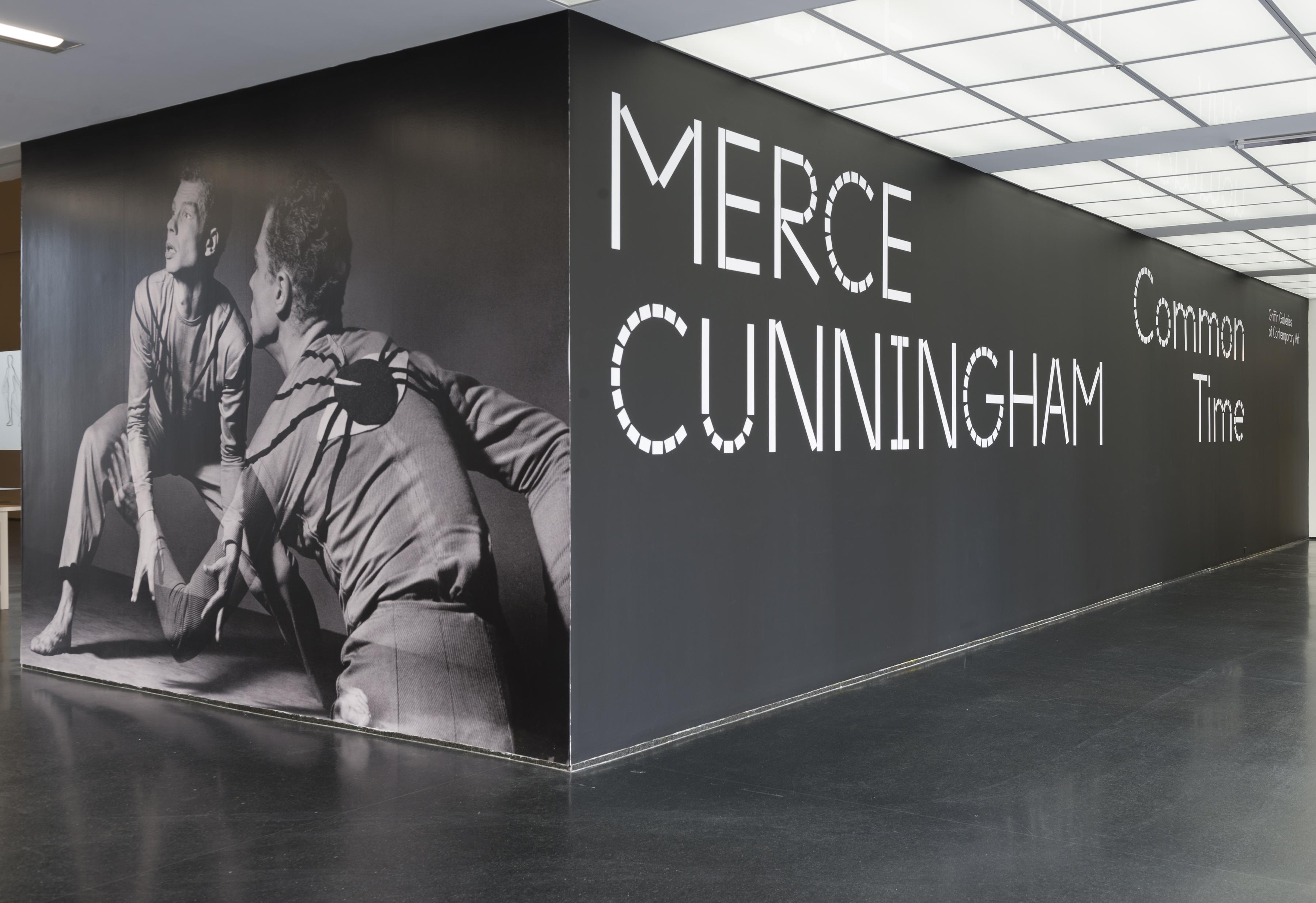 A black-walled corner in a room features a floor-to-ceiling gray-scale image of a crouching man seen from the front and the back on the left wall, and large white letters on the adjacent black wall that read: "MERCE CUNNINGHAM Common Time."