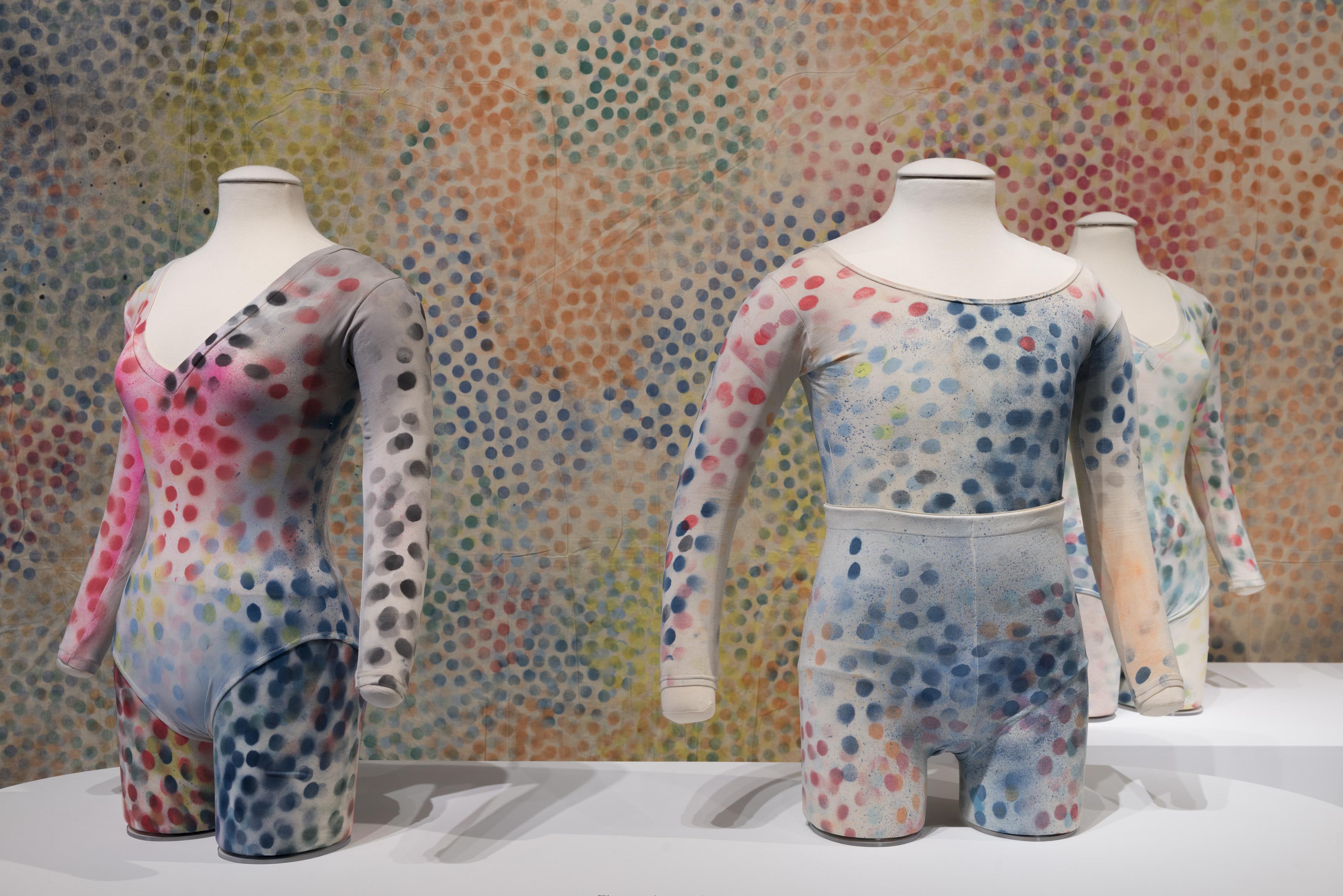 Two headless torso mannequins--one male one female--wear unitards covered in blue, red, and yellow dots, shown in front of a backdrop multicolored dots.