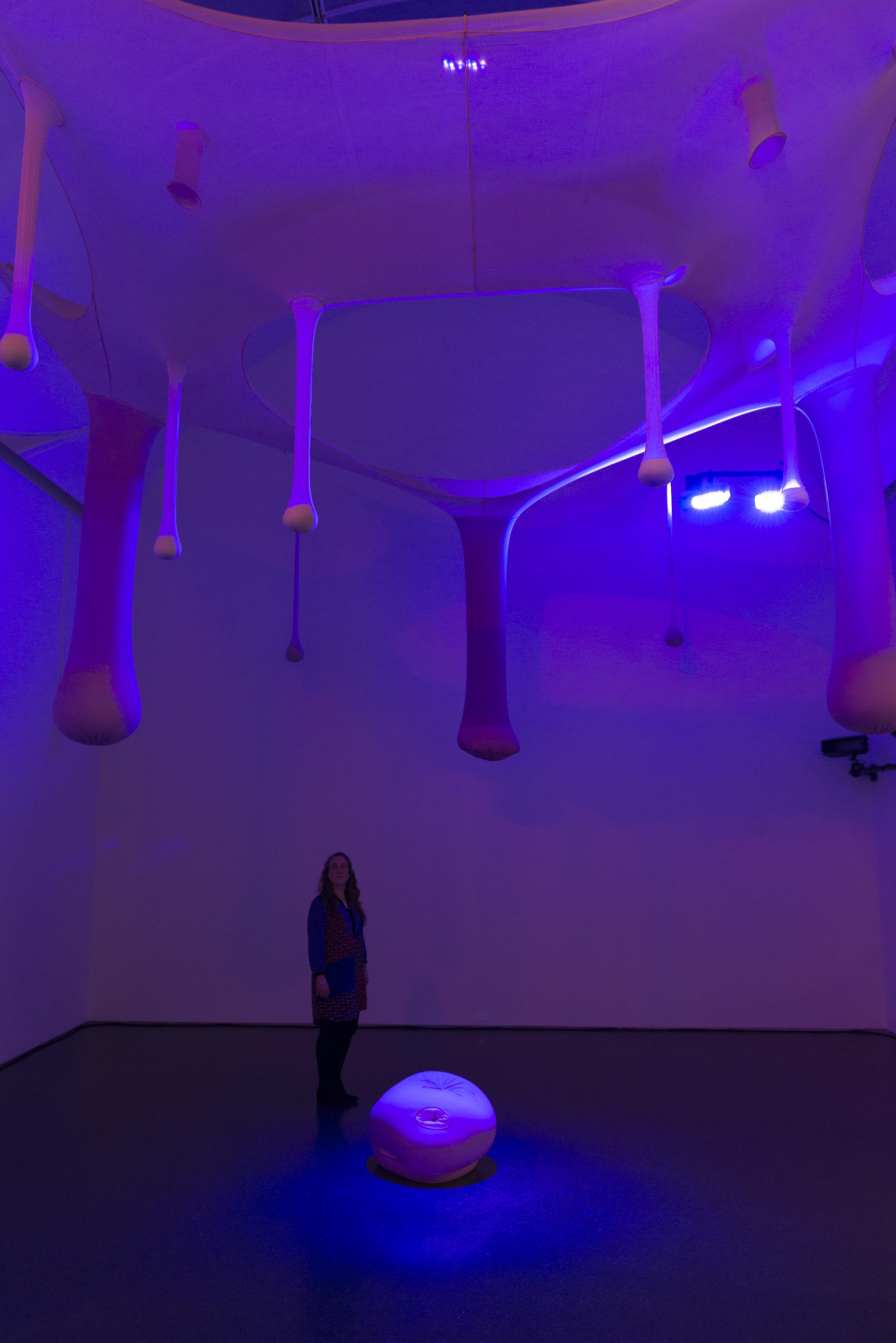 Organic oblong shapes hang from a suspended fabric in a room that glows purple and blue. A woman looks up at them, while standing next to a round purple object on the floor.