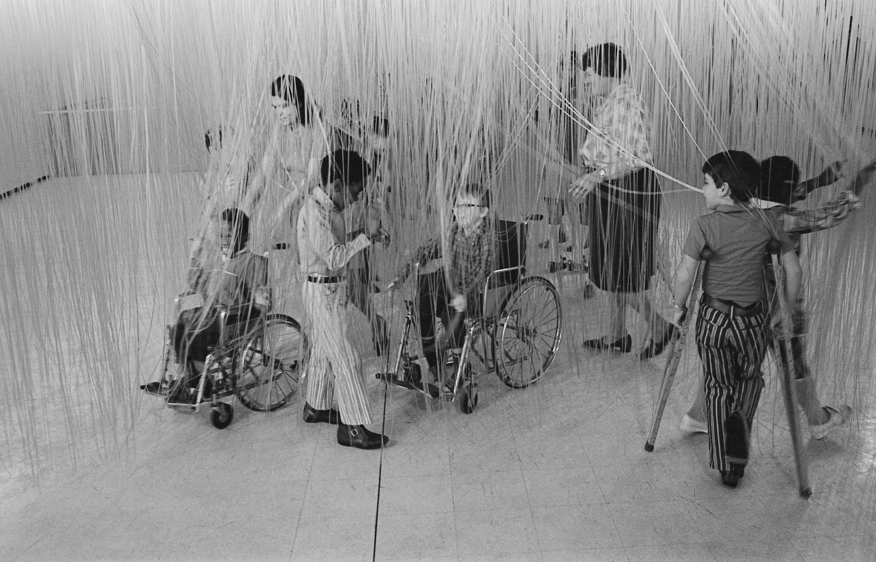 A group of children, some pushed in wheelchairs or using crutches, touches strands of an artwork hanging from the ceiling.