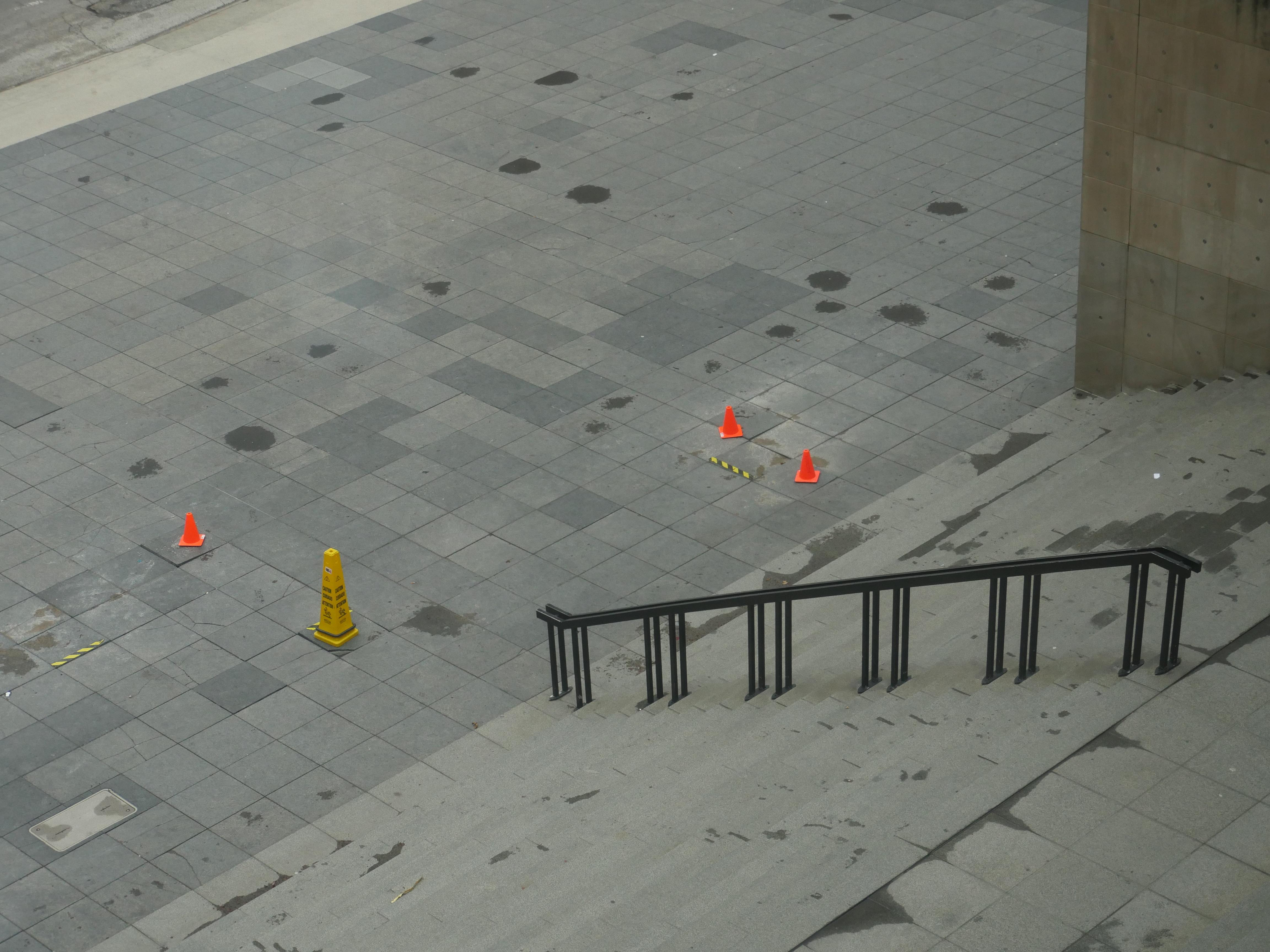 A stairway is shown from above, looking down on the plaza below where bright hazard cones dot the gray pavement tiles.