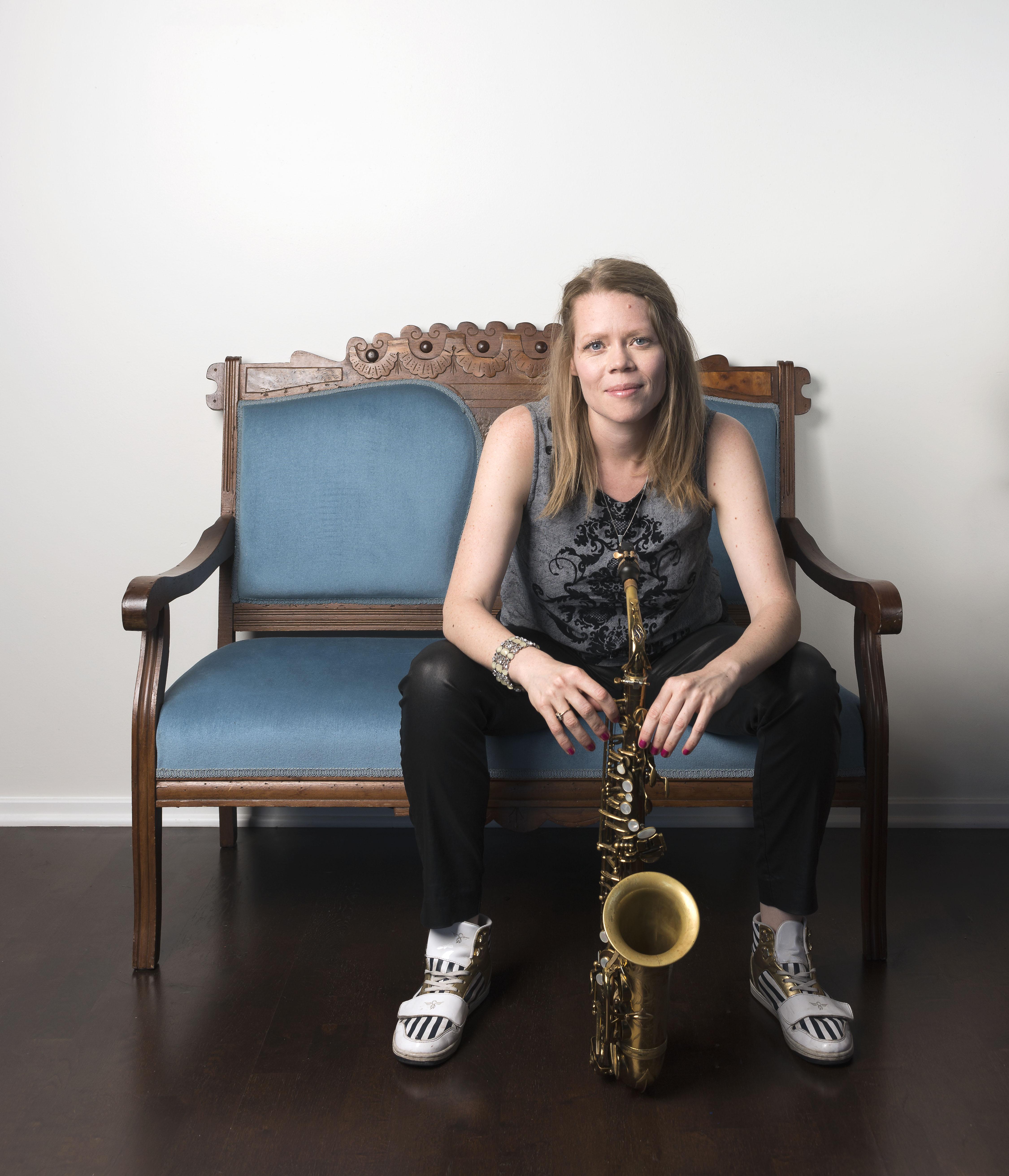 A blond, light-skinned woman sits facing forward and holding a saxophone that rests on the floor with both hands.