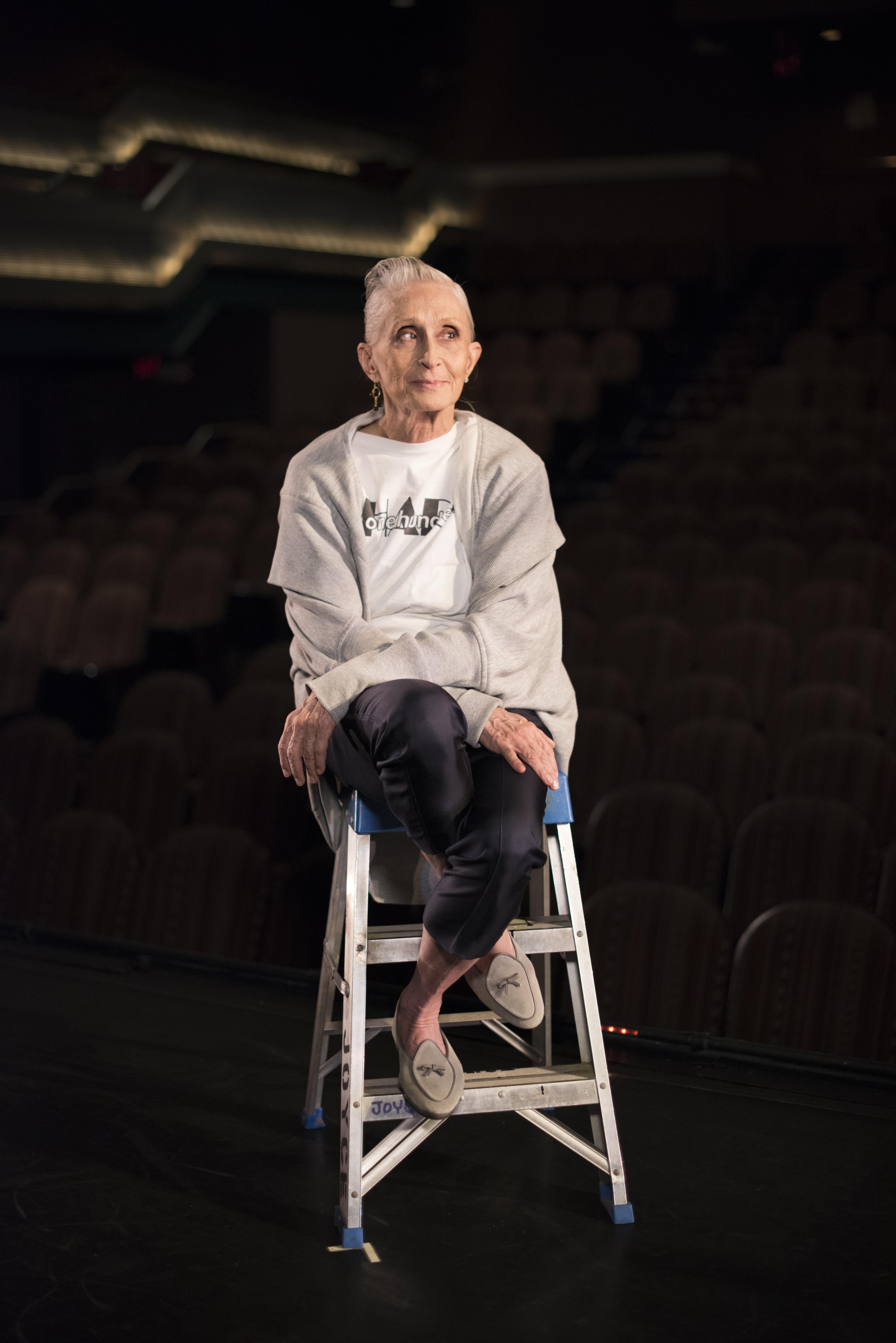 An older woman with her hair pulled back off her face, sits on top of a ladder on the edge of a stage with her legs crossed, as she looks to her left.